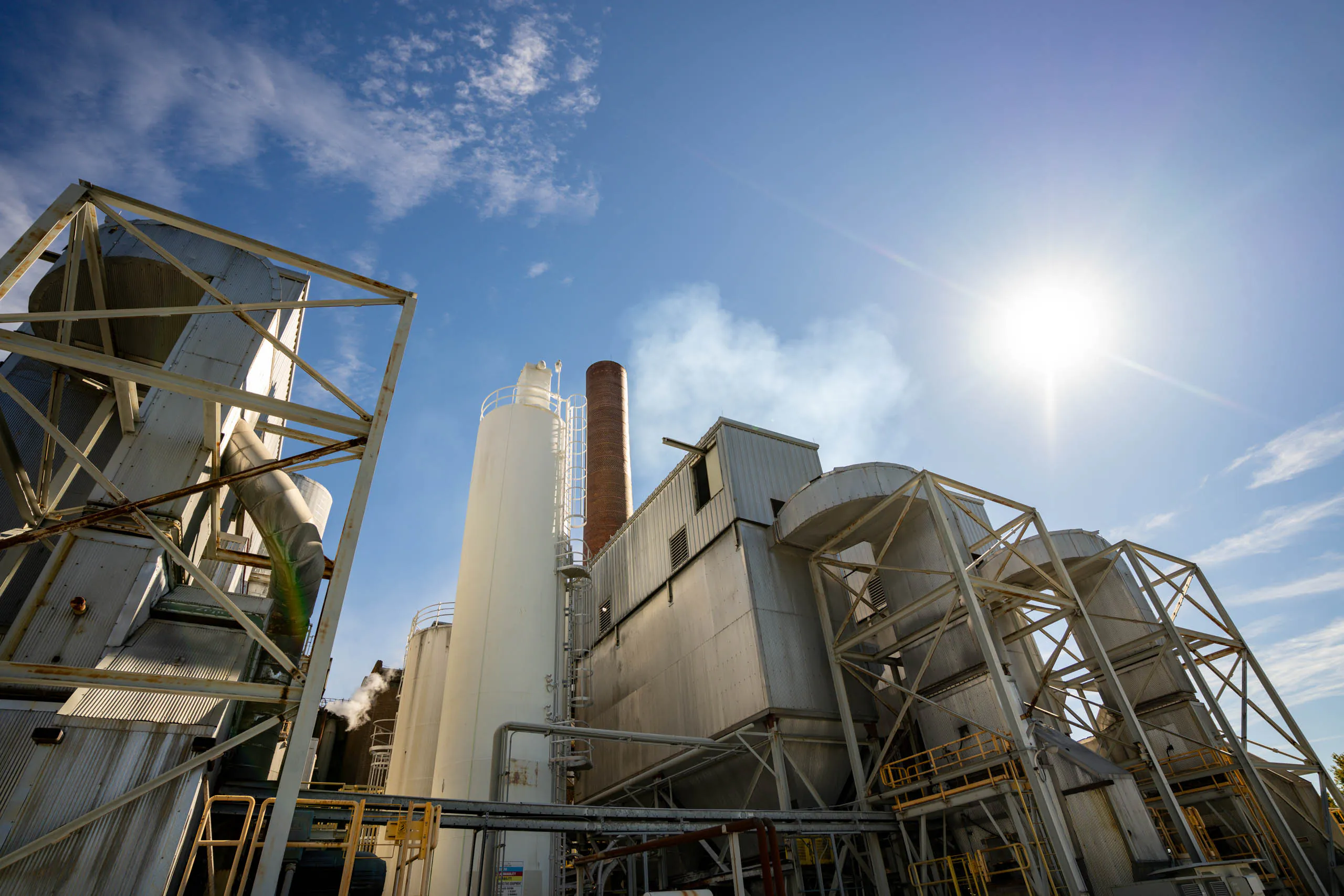 Large industrial factory with metal structures, silos, and smokestacks under a bright blue sky and sun. Some steam rises from the Lancaster Waste-To-Energy plant, highlighting its vital role in modern waste management. Ground-level perspective looking upward.