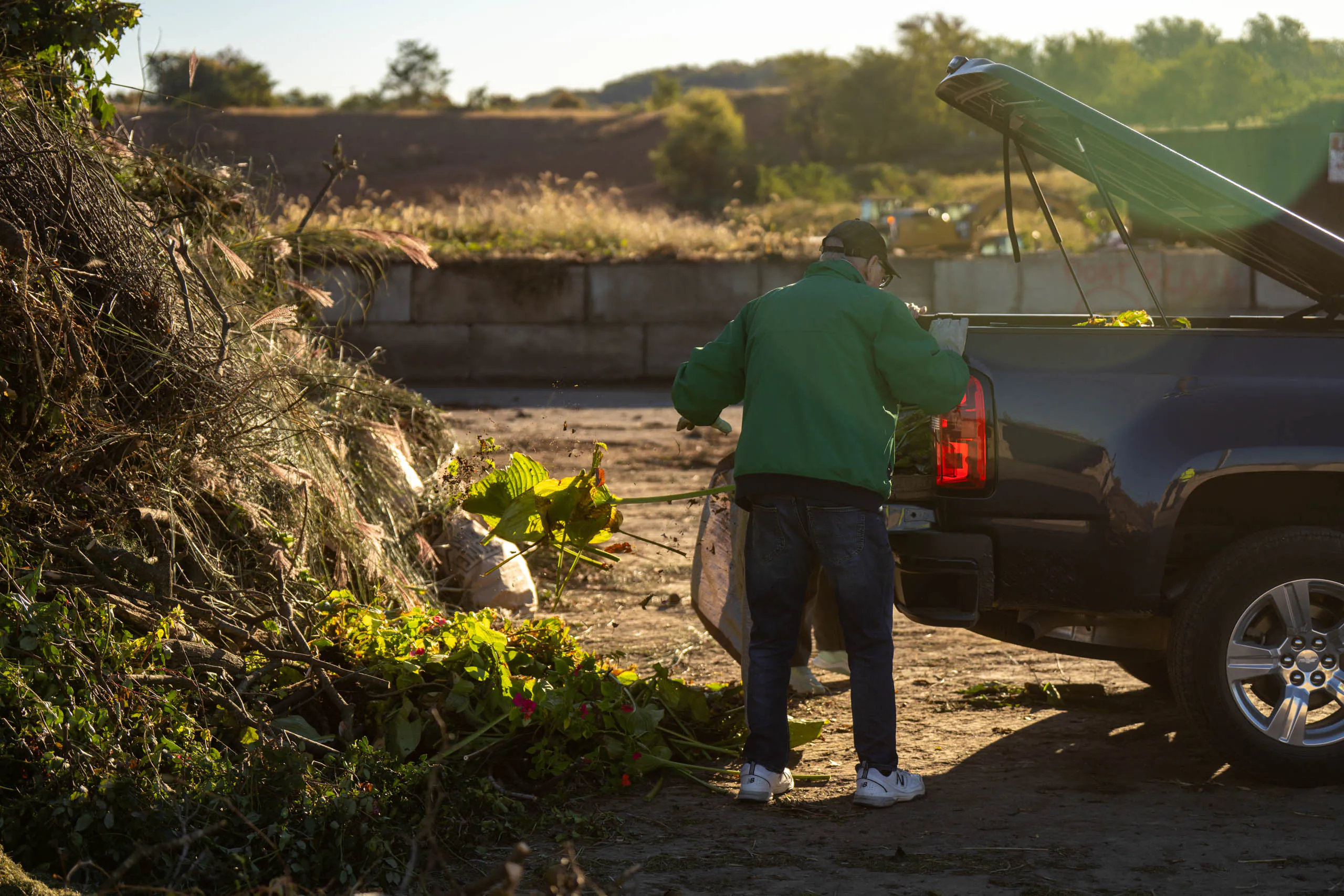 A person in a green jacket unloads yard waste from a pickup truck at the Frey Farm Landfill composting facility, with piles of branches and leaves nearby under bright sunlight.