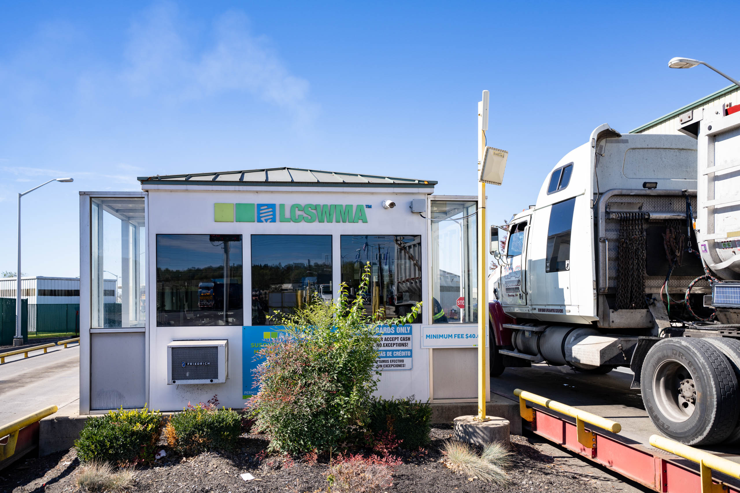 A small white booth with LCSWMA signage stands near a large white truck at the Susquehanna Resource Management Complex under a clear blue sky. Bushes and safety railings are in front of the booth.