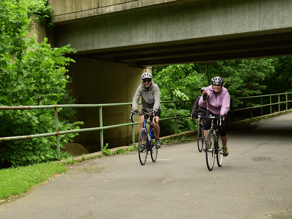 Two cyclists wearing helmets and jackets ride on a paved path under a concrete bridge, surrounded by green trees and metal railings. Both appear to be enjoying the ride.