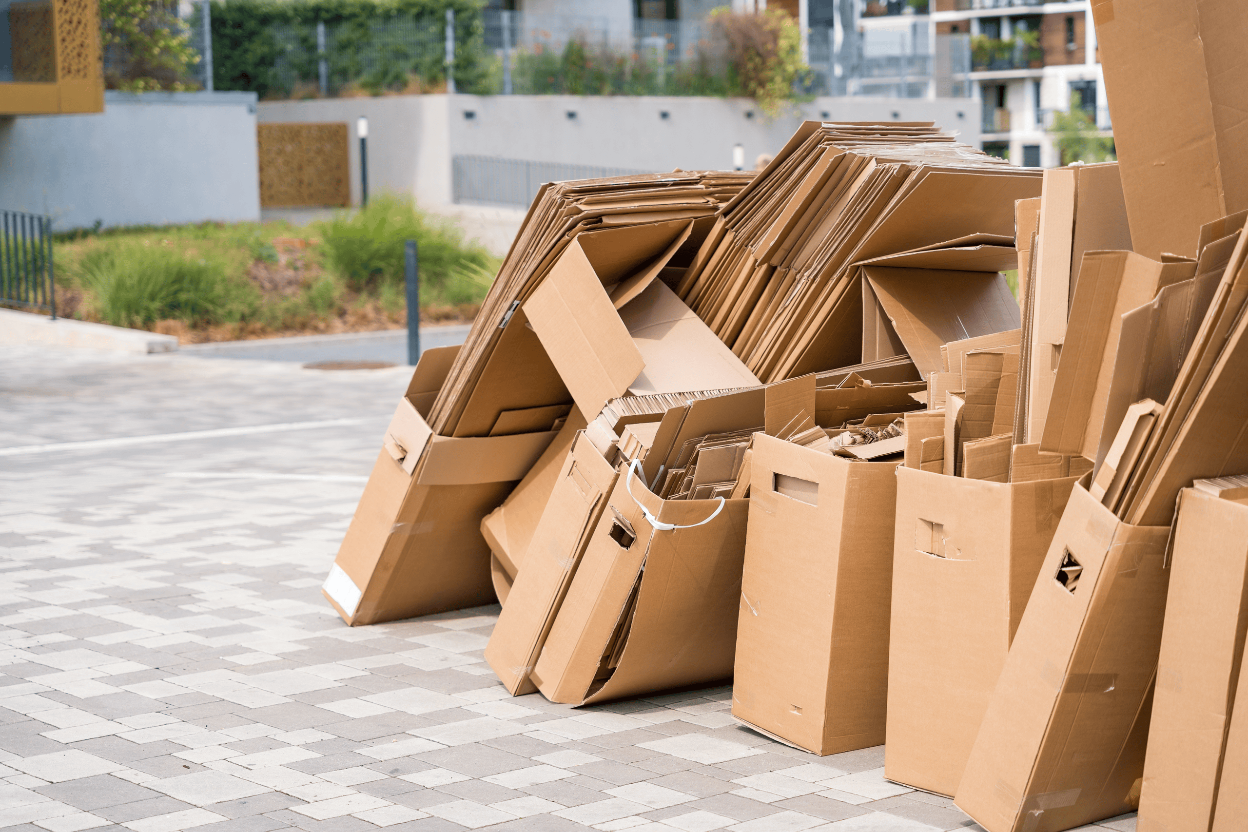 Stacks of flattened and open cardboard boxes are piled on a paved surface outdoors, inviting thoughts of Rethinking Waste in this residential or commercial area, with buildings and greenery in the background.