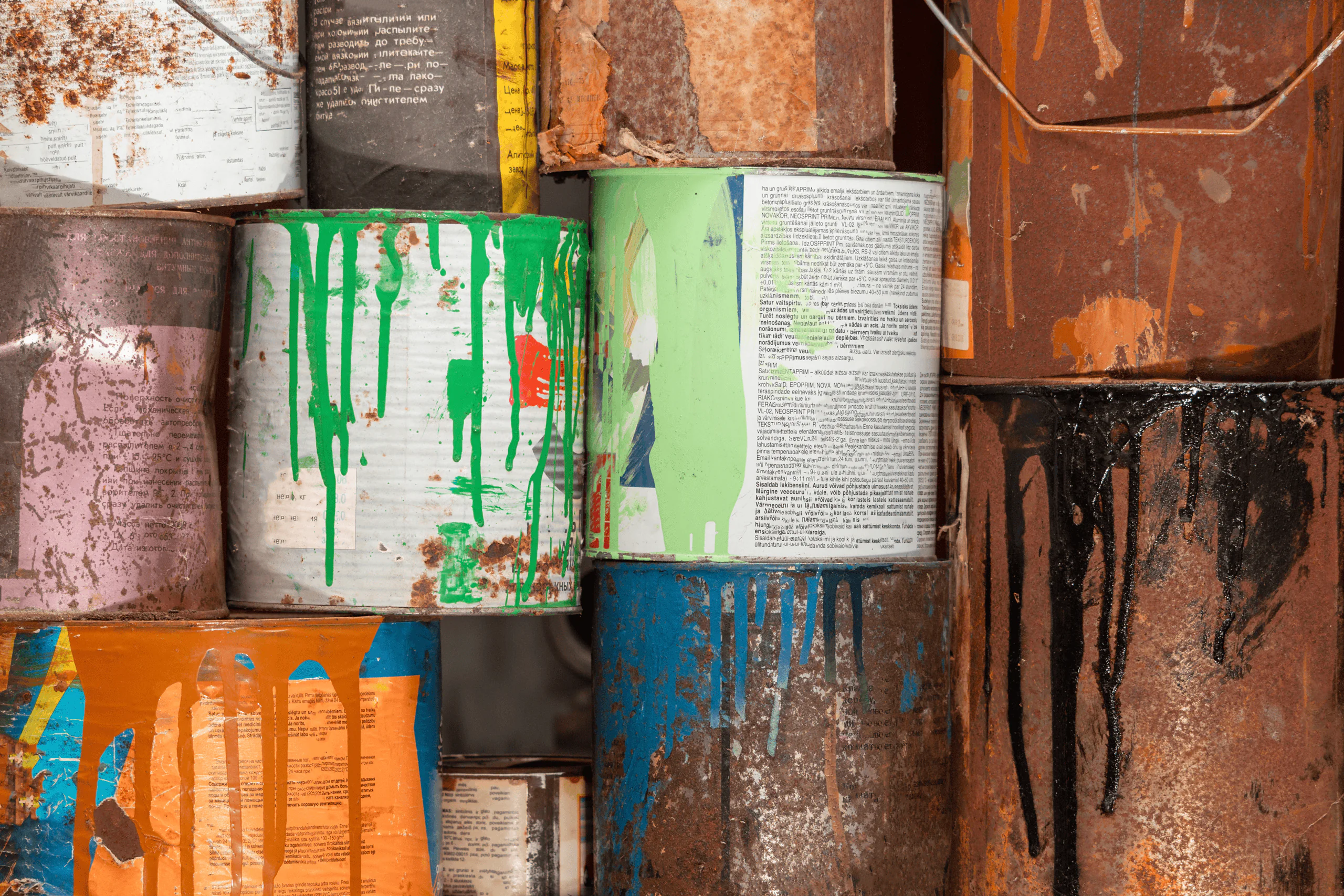 Stacks of old, rusty paint cans with colorful paint drips, including green, blue, orange, and black, running down their sides. The cans appear weathered and are tightly packed together.