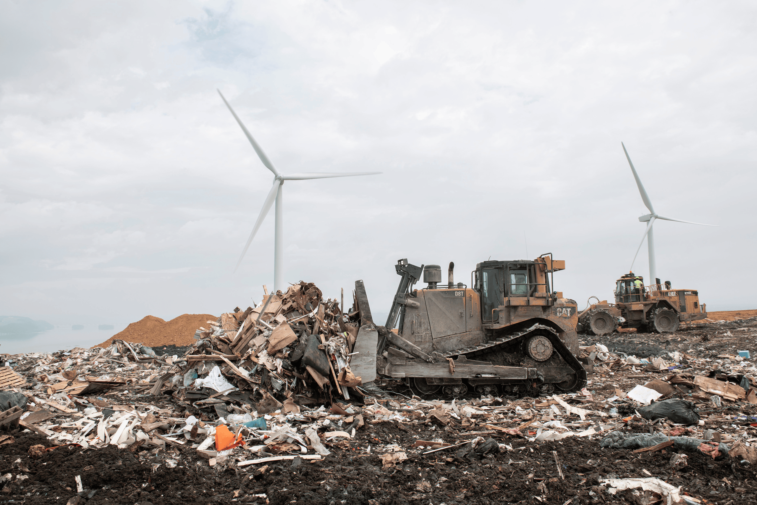 Bulldozers move piles of debris and waste at a lcswma site, showcasing waste management efforts, with wind turbines standing in the background against a cloudy sky.