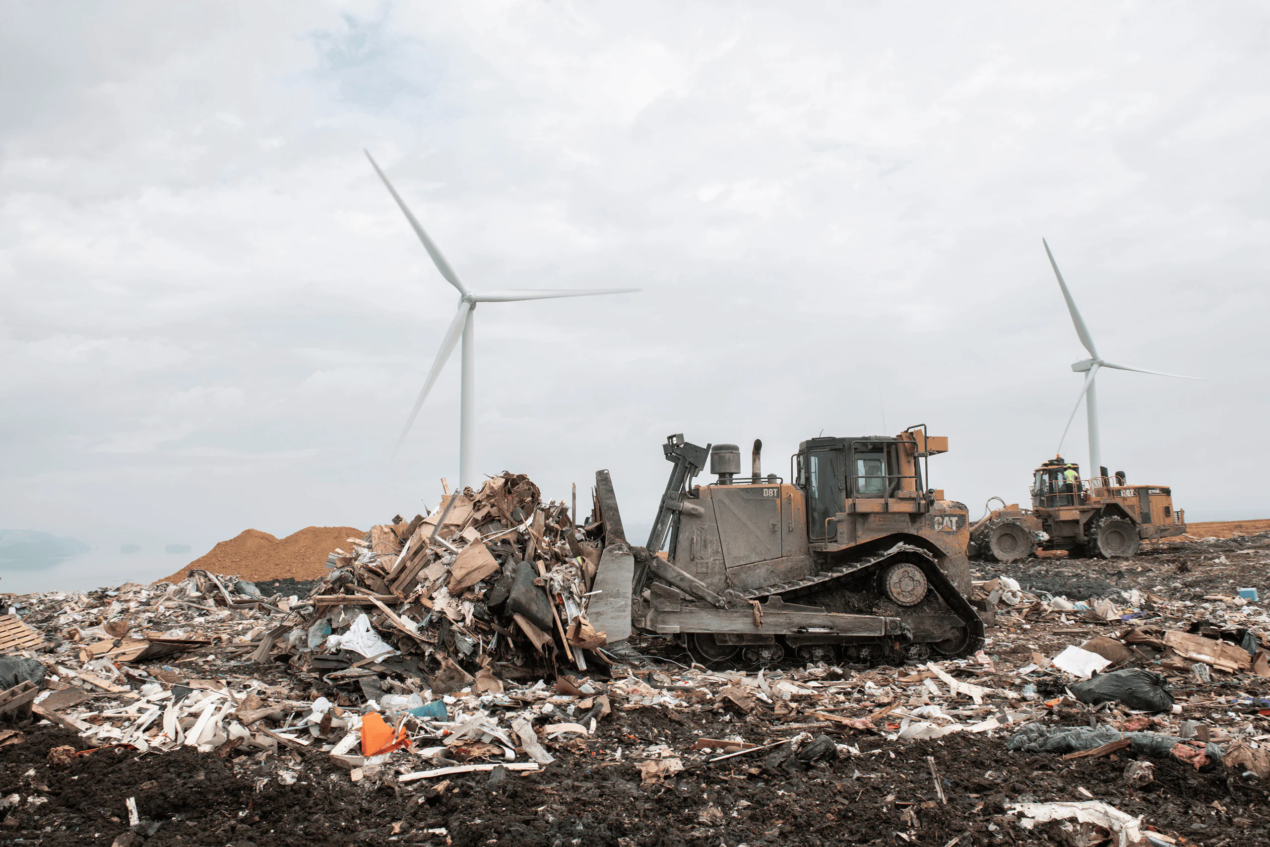 Bulldozers move piles of debris and waste at a lcswma site, showcasing waste management efforts, with wind turbines standing in the background against a cloudy sky.