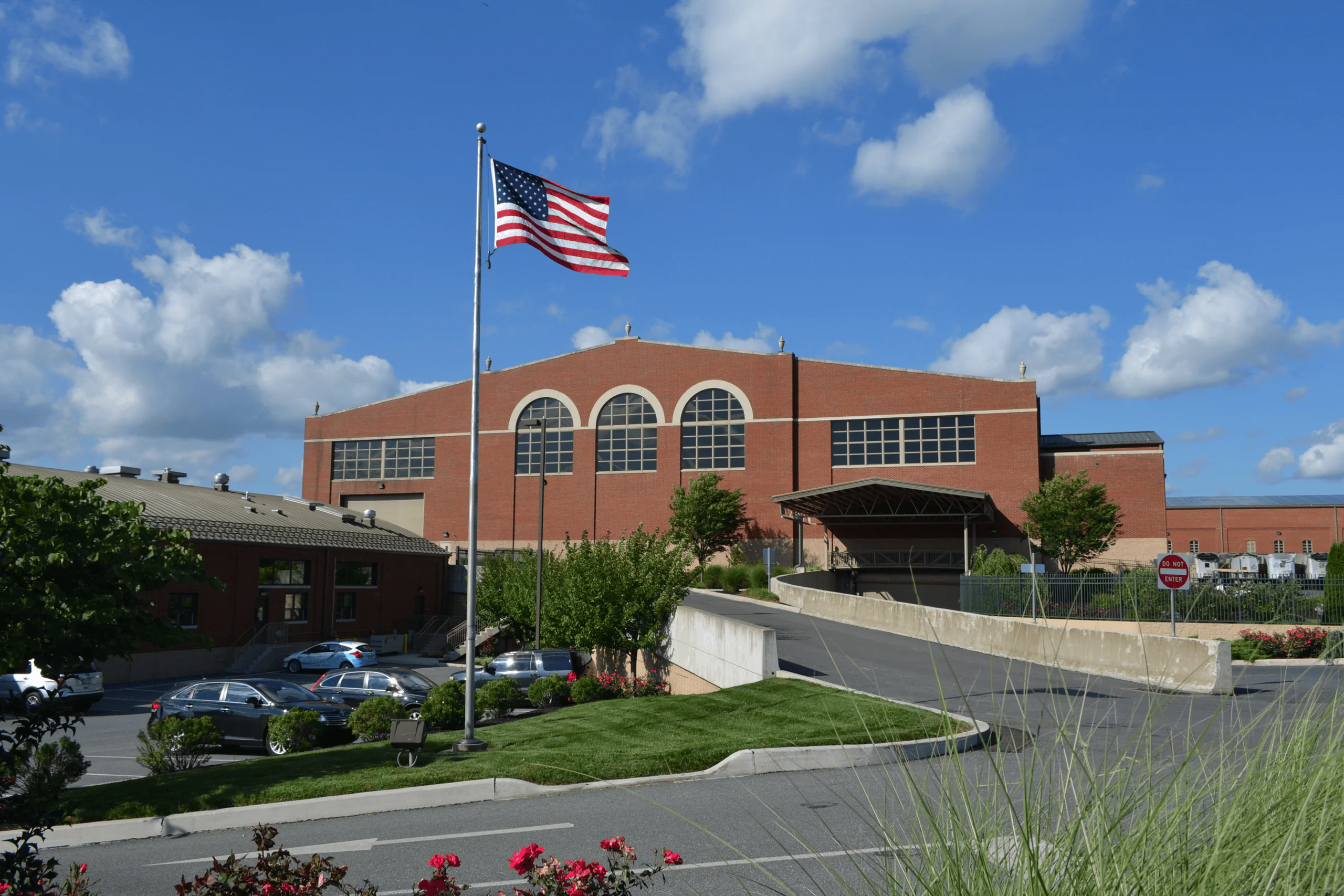 An American flag waves in front of a lcswma red brick building with arched windows and a covered entrance, surrounded by parked cars, greenery, and a clear blue sky with scattered clouds.