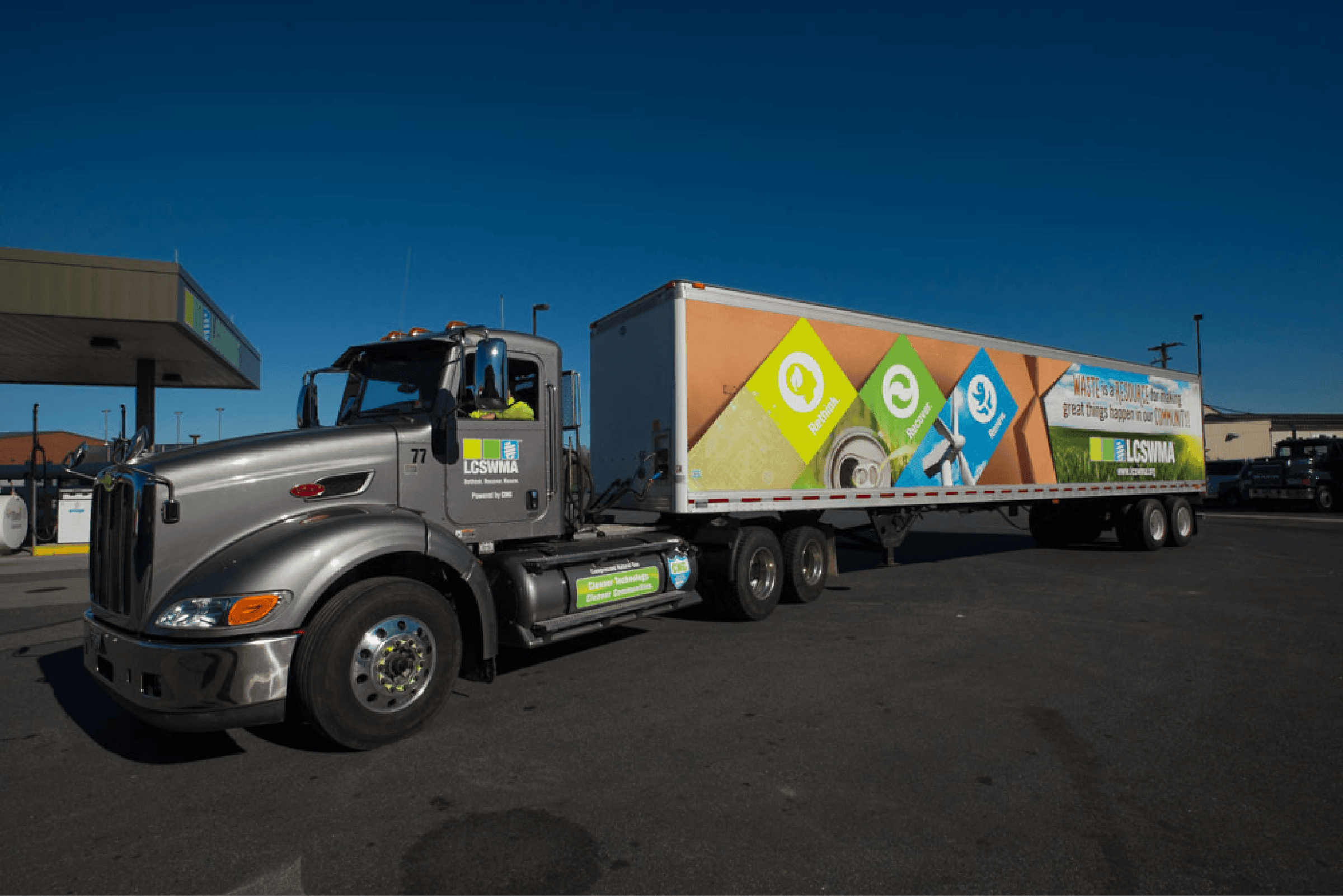 A large silver semi-truck with a colorful trailer is parked at lcswma station under a clear blue sky. The trailer displays eco-friendly advertisements, rethinking waste with green and blue graphics and text.