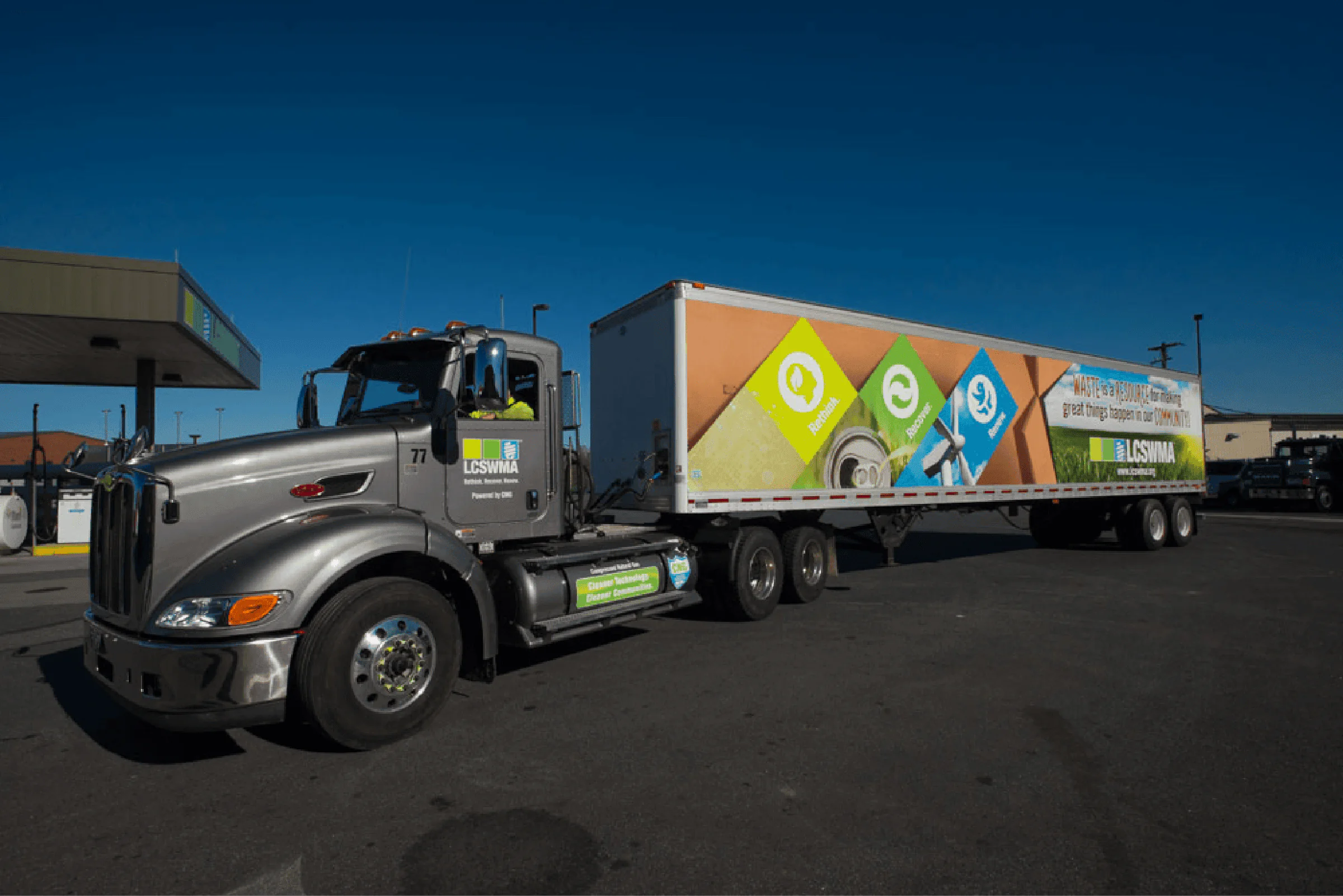 A large silver semi-truck with a colorful trailer is parked at lcswma station under a clear blue sky. The trailer displays eco-friendly advertisements, rethinking waste with green and blue graphics and text.