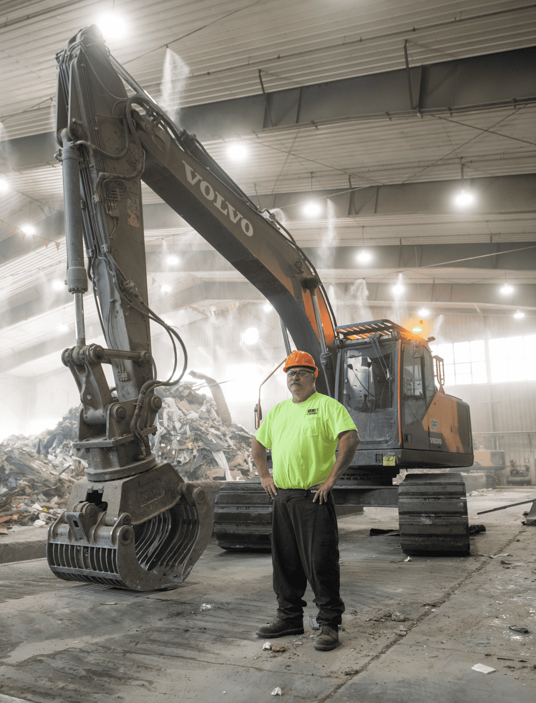 A lcswma worker in a neon yellow shirt and hard hat stands in front of a large Volvo excavator inside an industrial building filled with debris.