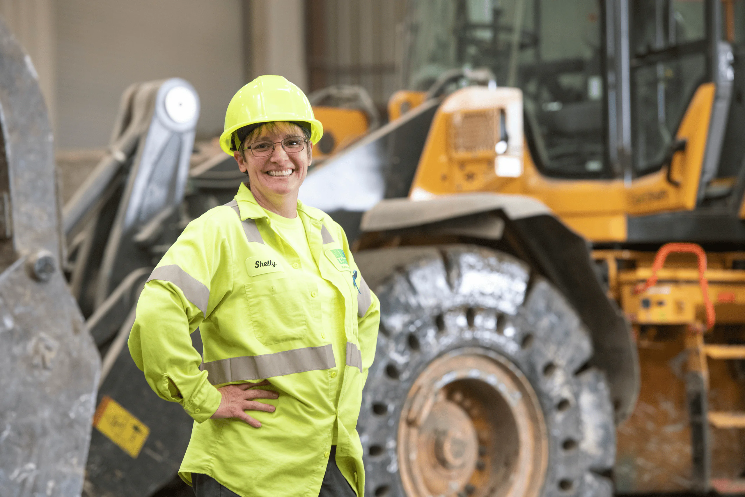 A woman wearing a yellow hard hat and high-visibility jacket smiles while standing in front of a large lcswma vehicle inside an industrial building.