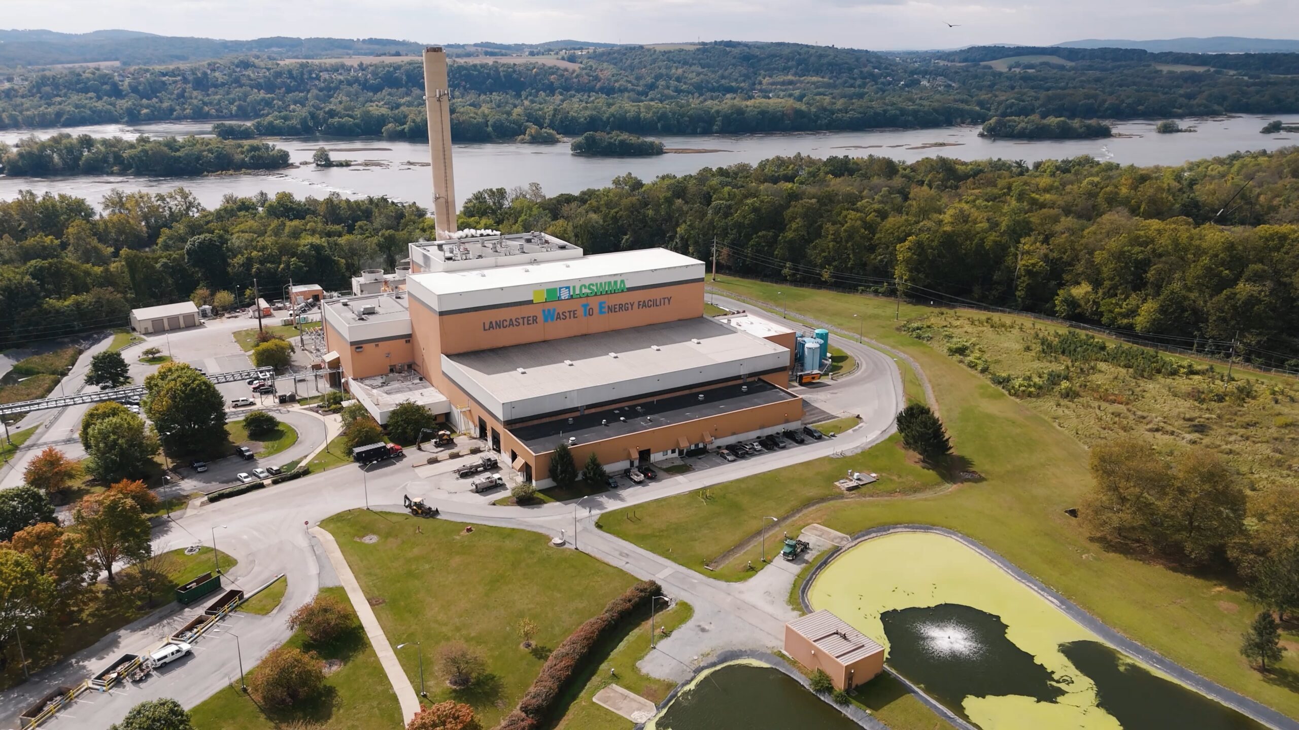 Aerial view of the Lancaster Waste-To-Energy Facility, a key waste management energy facility surrounded by trees, with a river in the background and small ponds in the foreground.