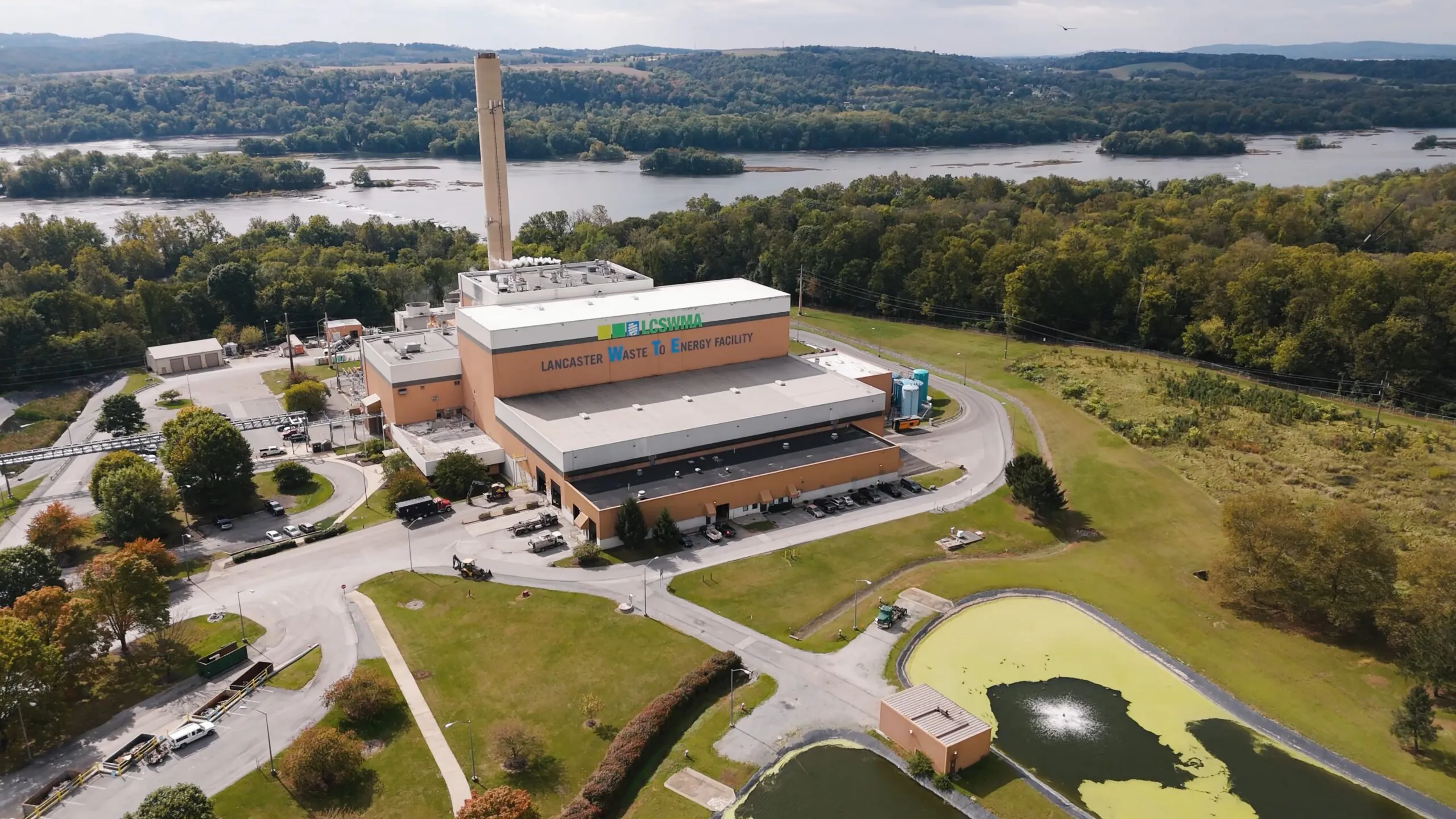 Aerial view of the Lancaster Waste-To-Energy Facility, a key waste management energy facility surrounded by trees, with a river in the background and small ponds in the foreground.
