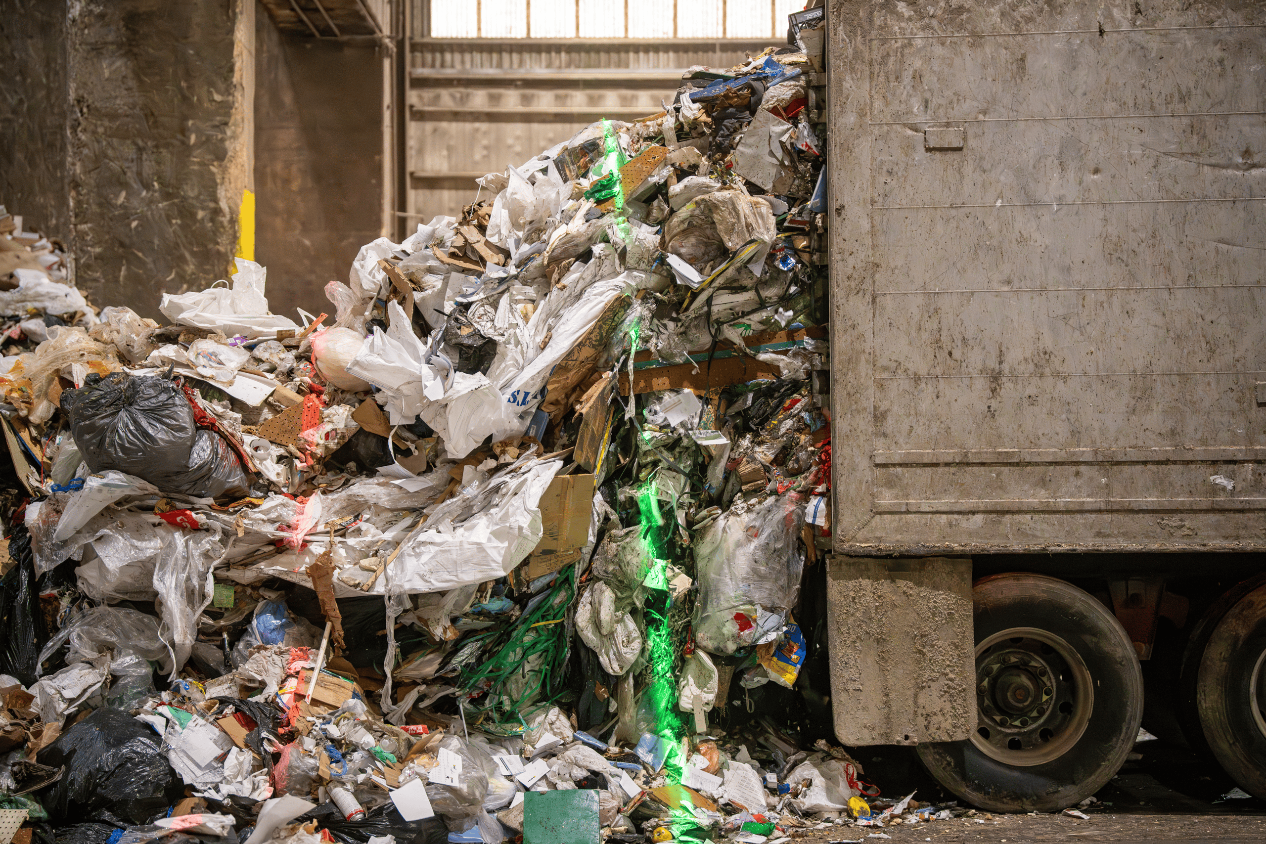 A large pile of mixed garbage and recyclables spills out of the back of a truck inside an industrial waste facility, with a bright green line of light running through the trash.