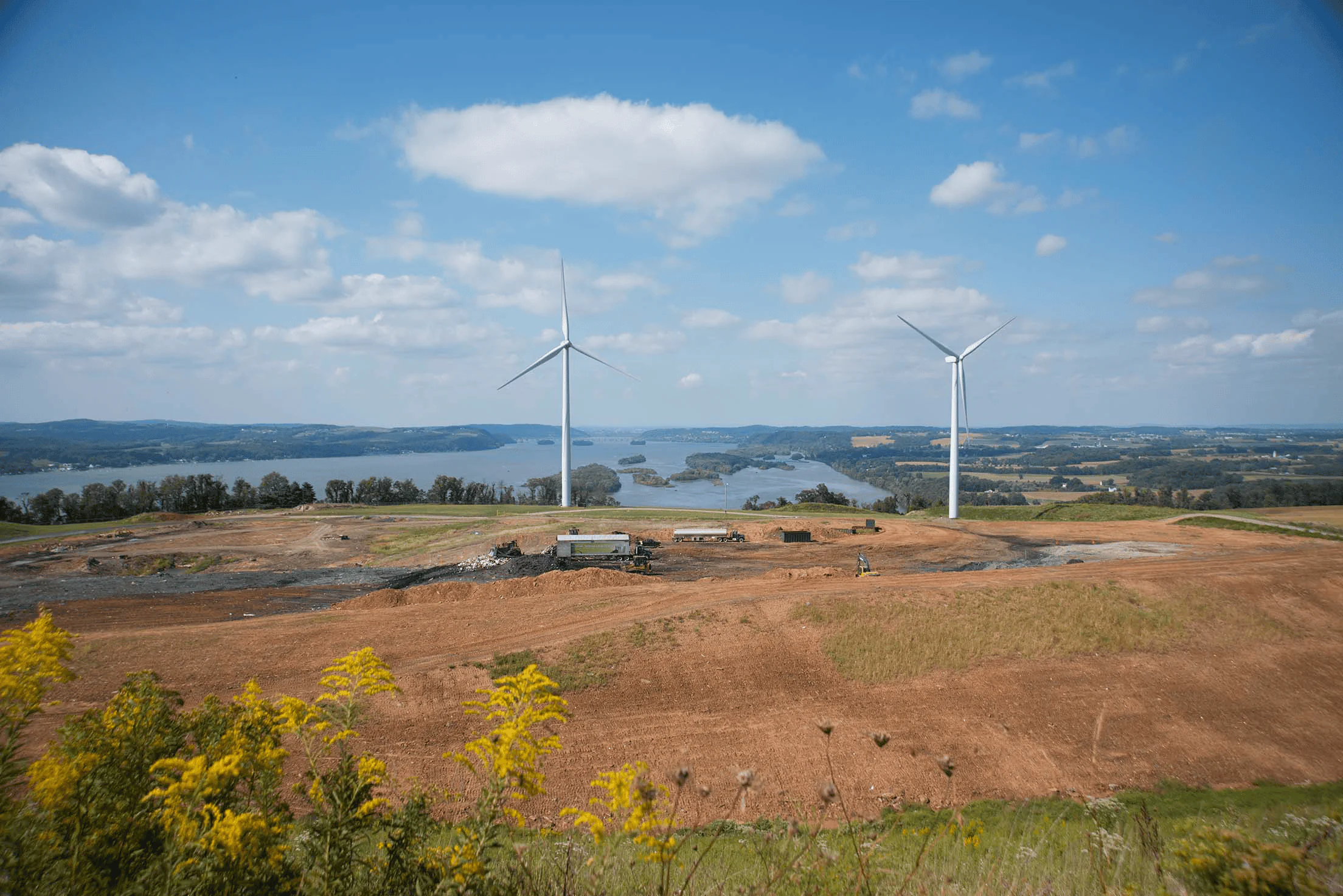 Two large wind turbines stand on a grassy hill overlooking a river and distant hills, with a partly cloudy sky above. Construction vehicles and cleared land are visible in the foreground.