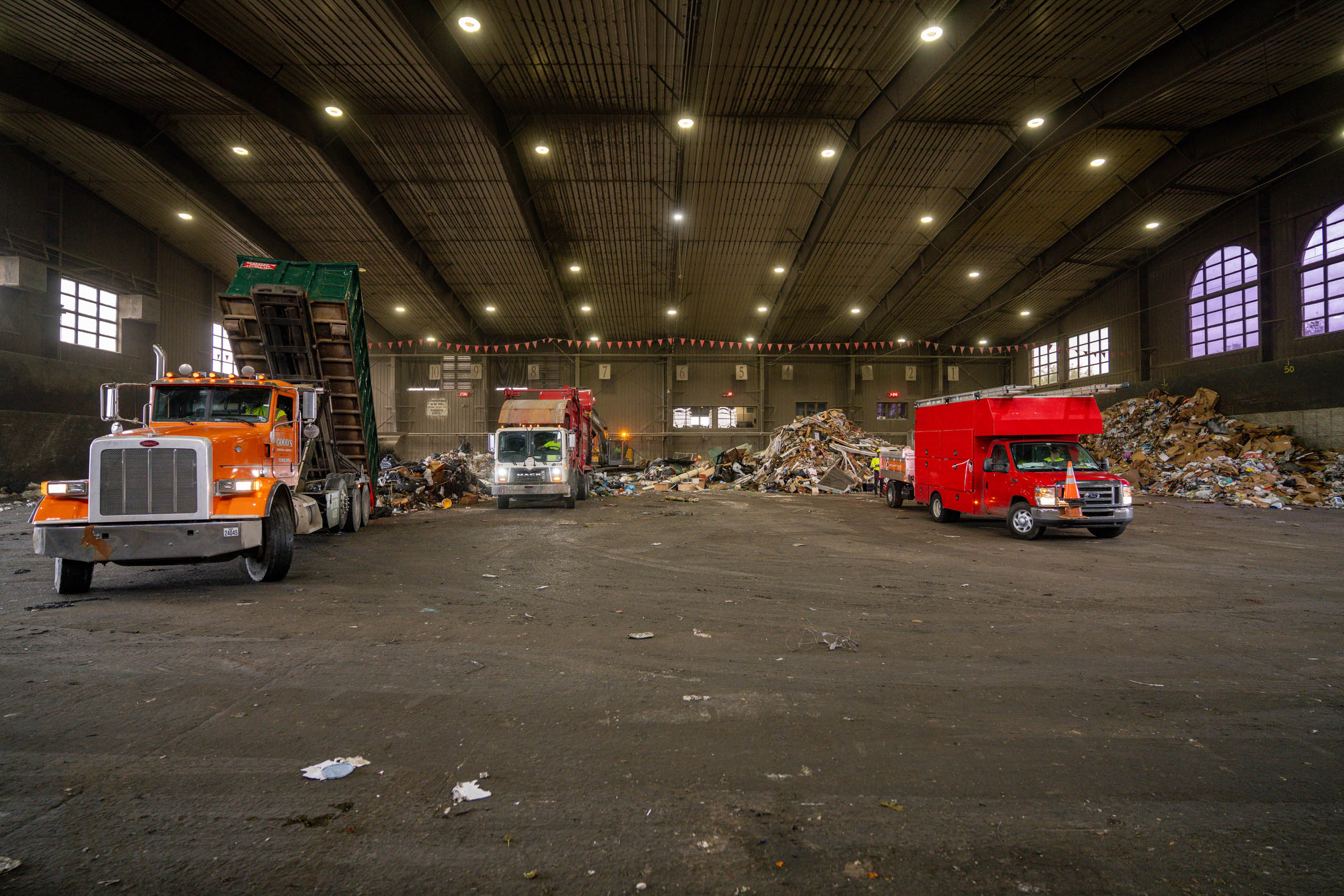 Three trucks, including a garbage truck and two red utility vehicles, are parked inside a large lcswma warehouse filled with piles of trash and debris under bright overhead lights.