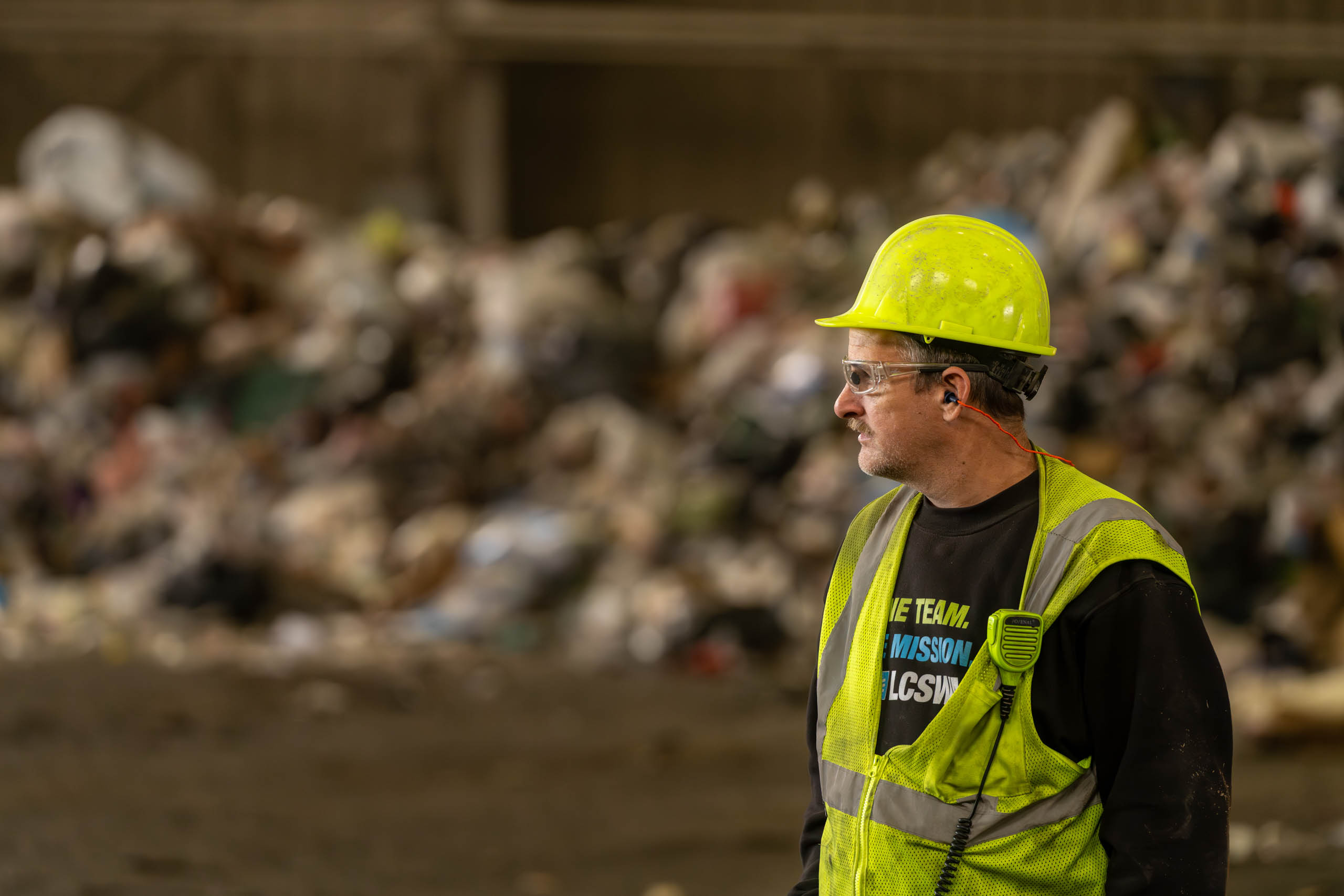 A lcswma worker wearing a yellow hard hat, safety glasses, and a reflective vest stands in front of a large pile of garbage or recyclables, looking to the side in an industrial setting.