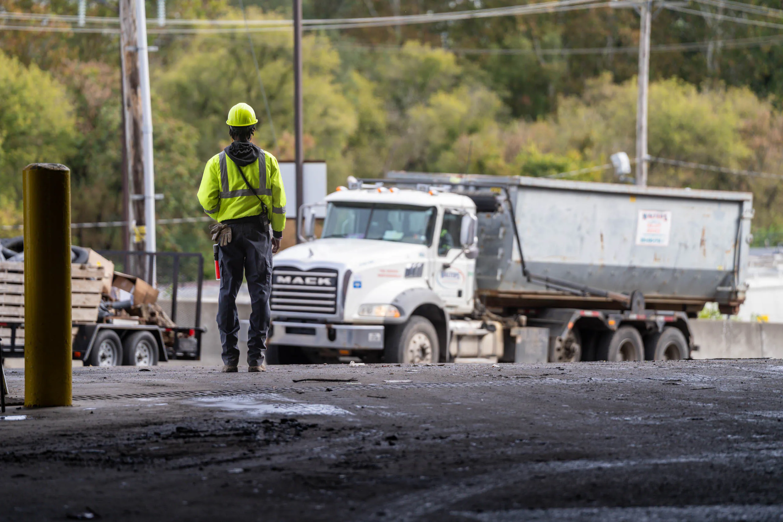 A lcswma worker in a yellow safety vest and helmet stands facing a white dump truck on a worksite, where Residential Disposal Rates influence the project's efficiency, with trees and power lines seen in the background.