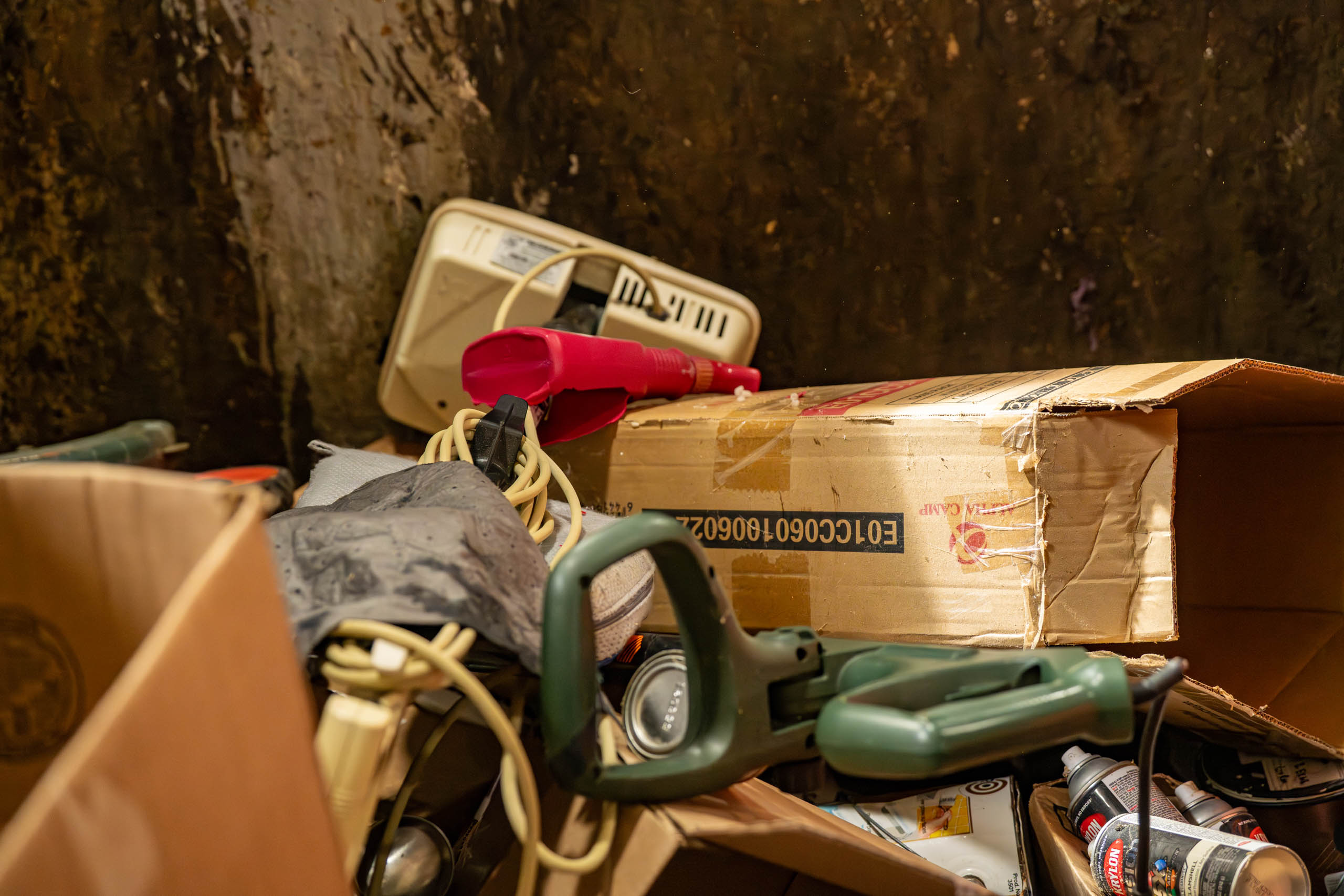 A pile of assorted discarded items in cardboard boxes, including tangled electrical cords, a red appliance, and a scale sits against a dirty, stained wall—reminding you to Plan Your Drop-off for responsible disposal.