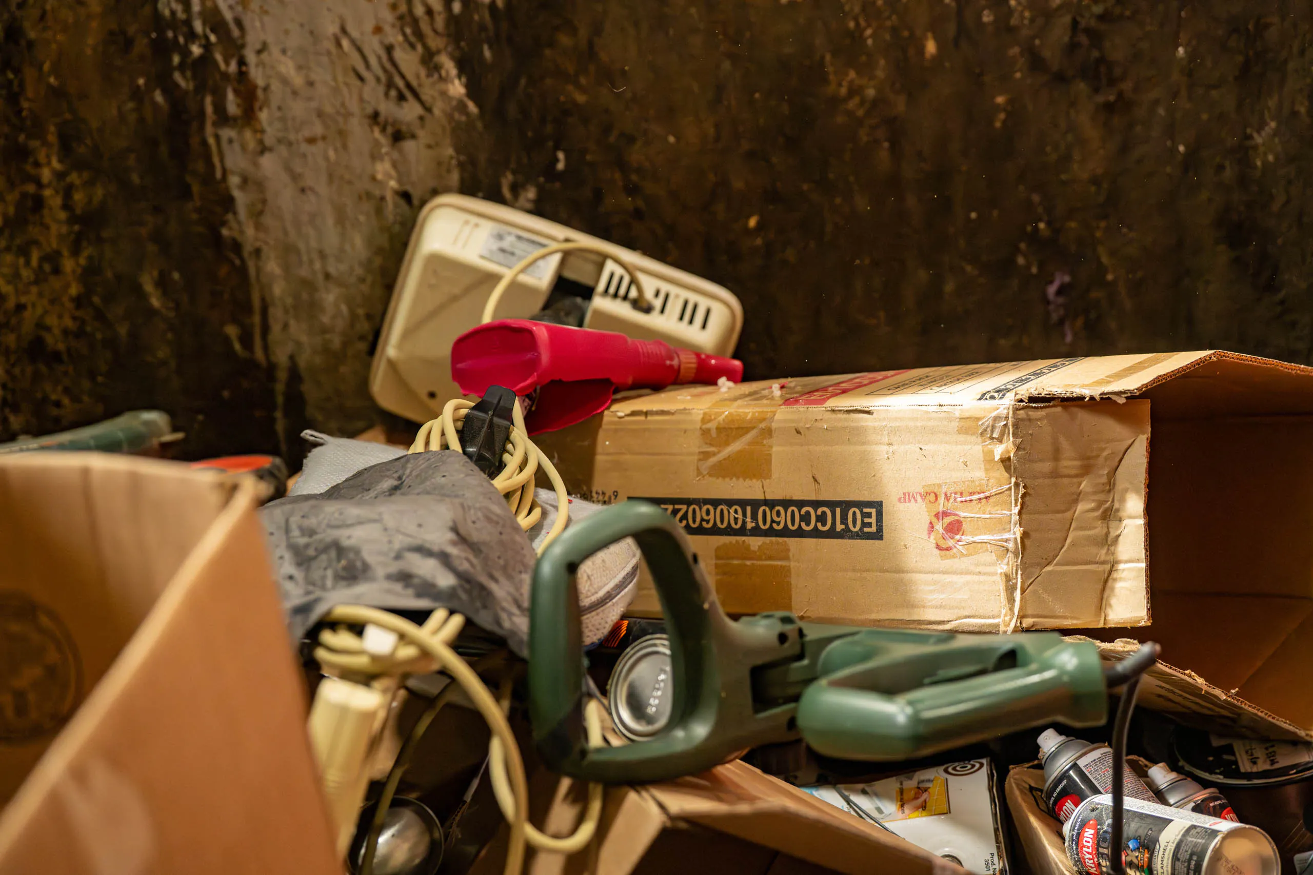 A pile of assorted discarded items in cardboard boxes, including tangled electrical cords, a red appliance, and a scale sits against a dirty, stained wall—reminding you to Plan Your Drop-off for responsible disposal.