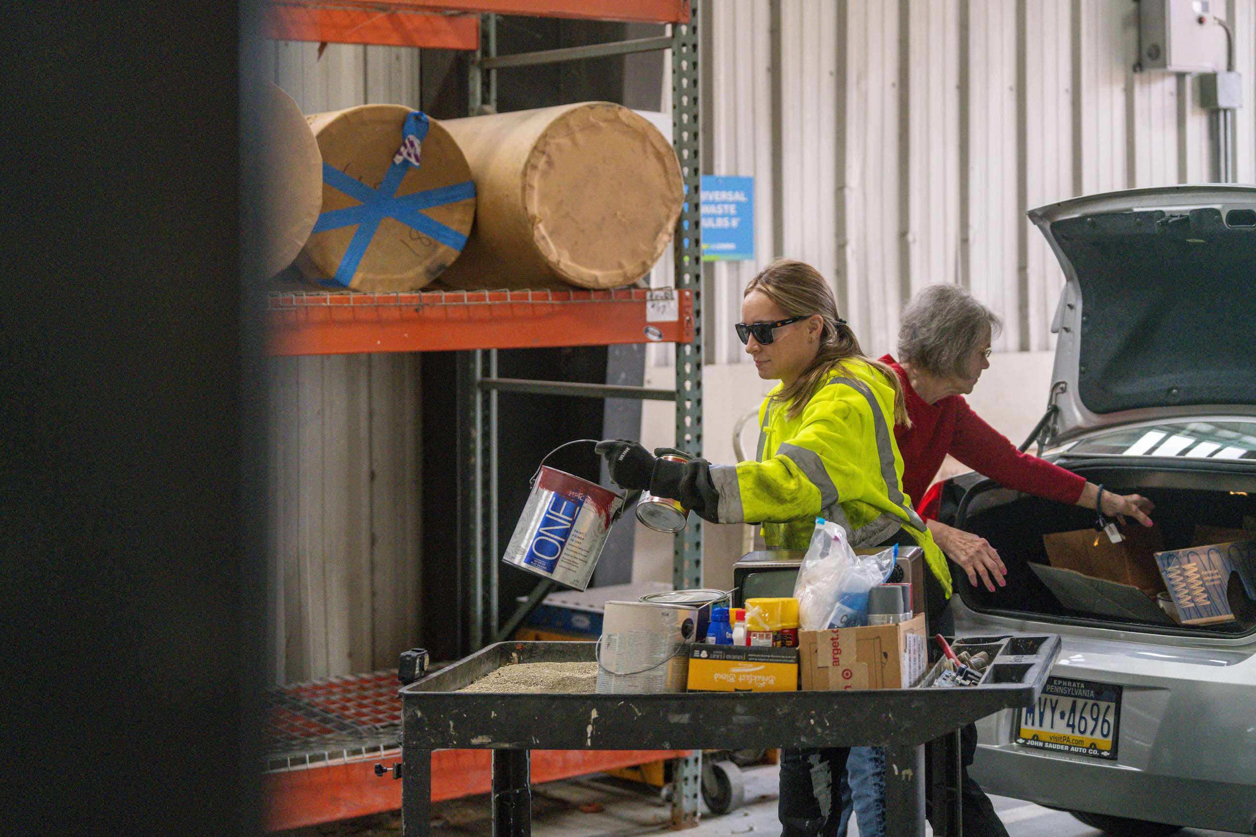 A lcswma woman in a yellow safety jacket and sunglasses pours paint into a container at a recycling center, while another woman unloads items from the trunk of a car nearby. Shelves and recycling materials are visible in the background.
