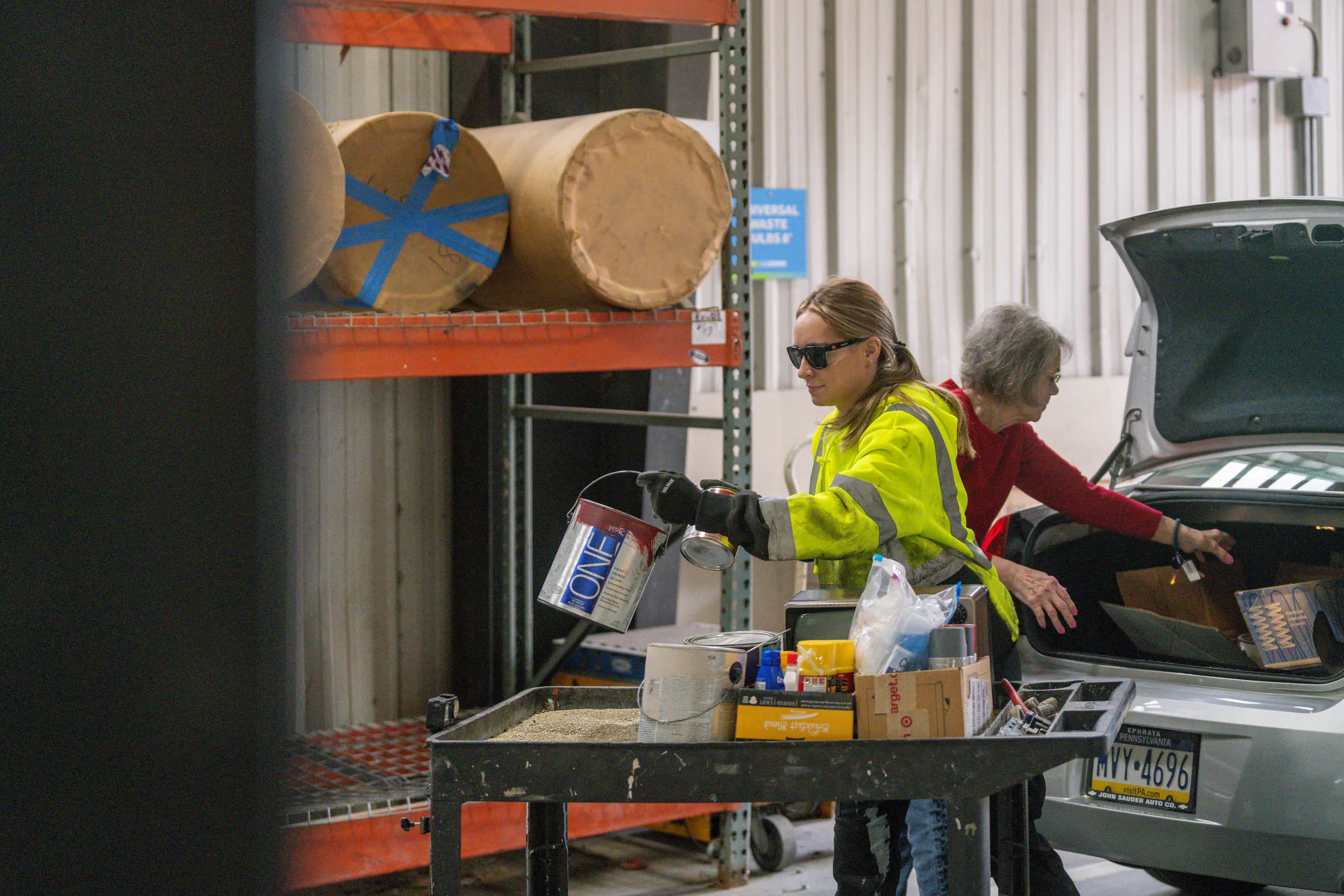 A lcswma woman in a yellow safety jacket and sunglasses pours paint into a container at a recycling center, while another woman unloads items from the trunk of a car nearby. Shelves and recycling materials are visible in the background.
