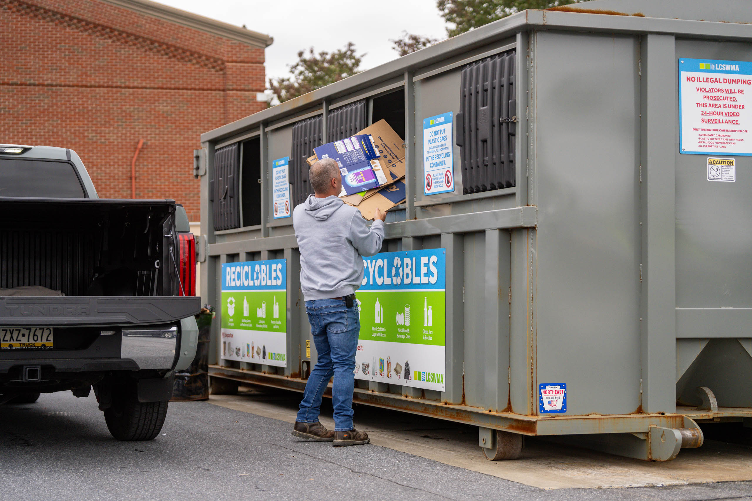A person uses drop-off planning as they place flattened cardboard boxes into a large outdoor recycling dumpster next to a parked black pickup truck. The dumpster features clear signs and labels for recyclables.