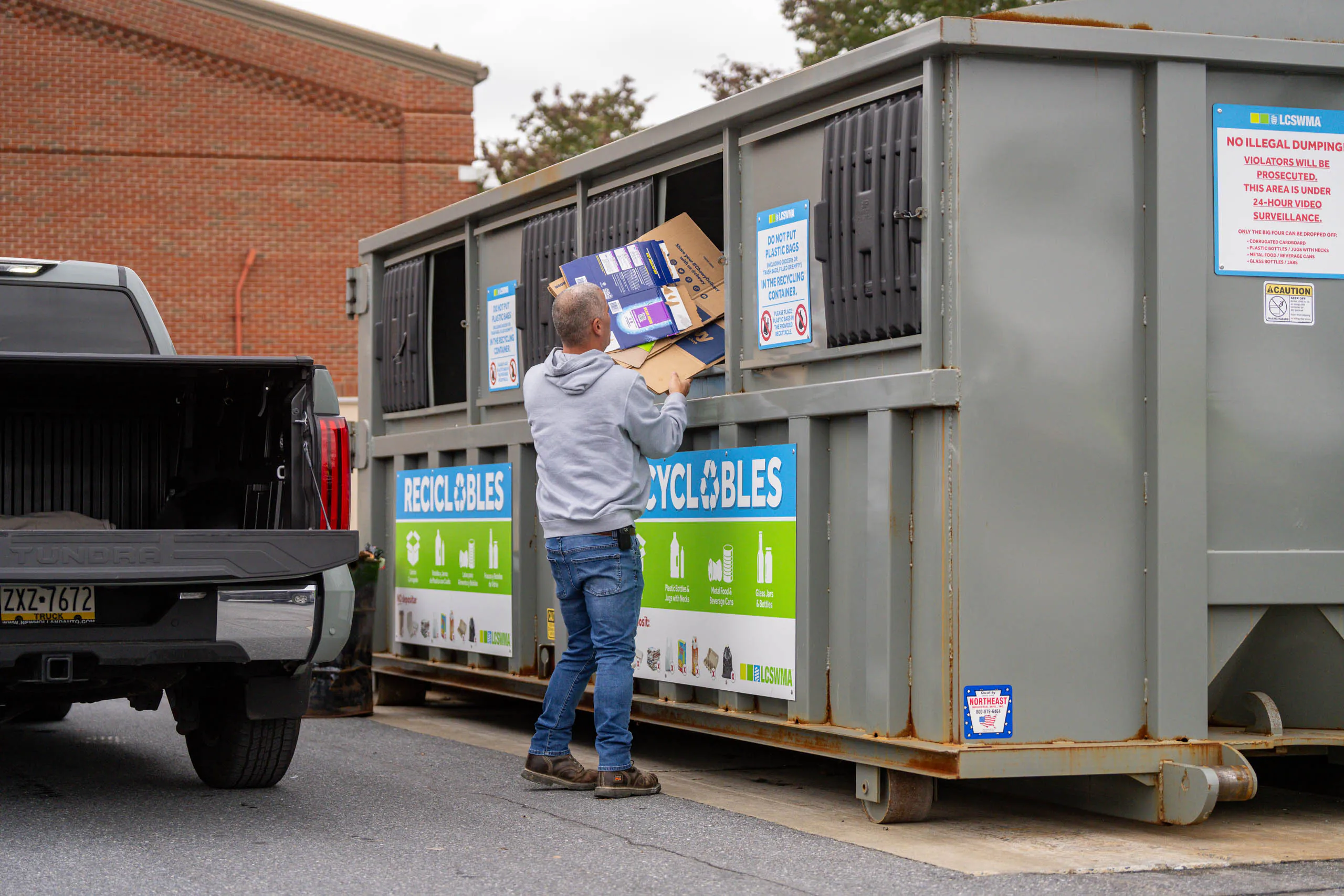 A person uses drop-off planning as they place flattened cardboard boxes into a large outdoor recycling dumpster next to a parked black pickup truck. The dumpster features clear signs and labels for recyclables.