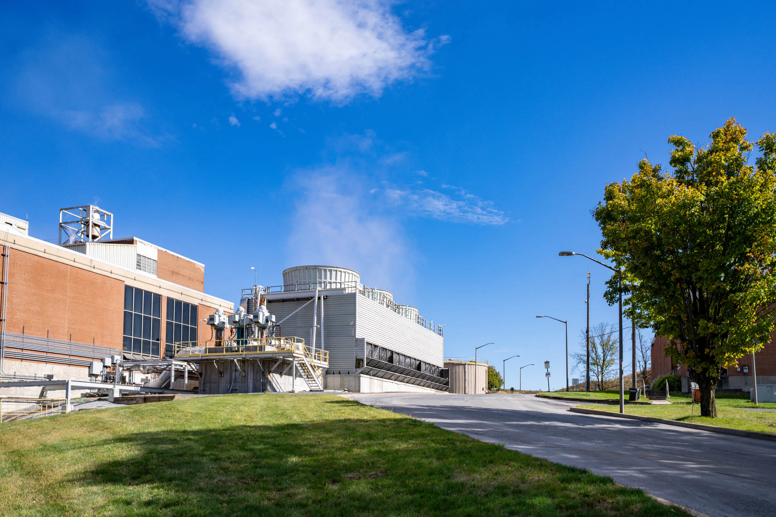 A large lcswma building with brick and metal structures, cooling towers emitting steam, and a clear blue sky. Green grass and a leafy tree are in the foreground, with a paved road leading alongside the facility.