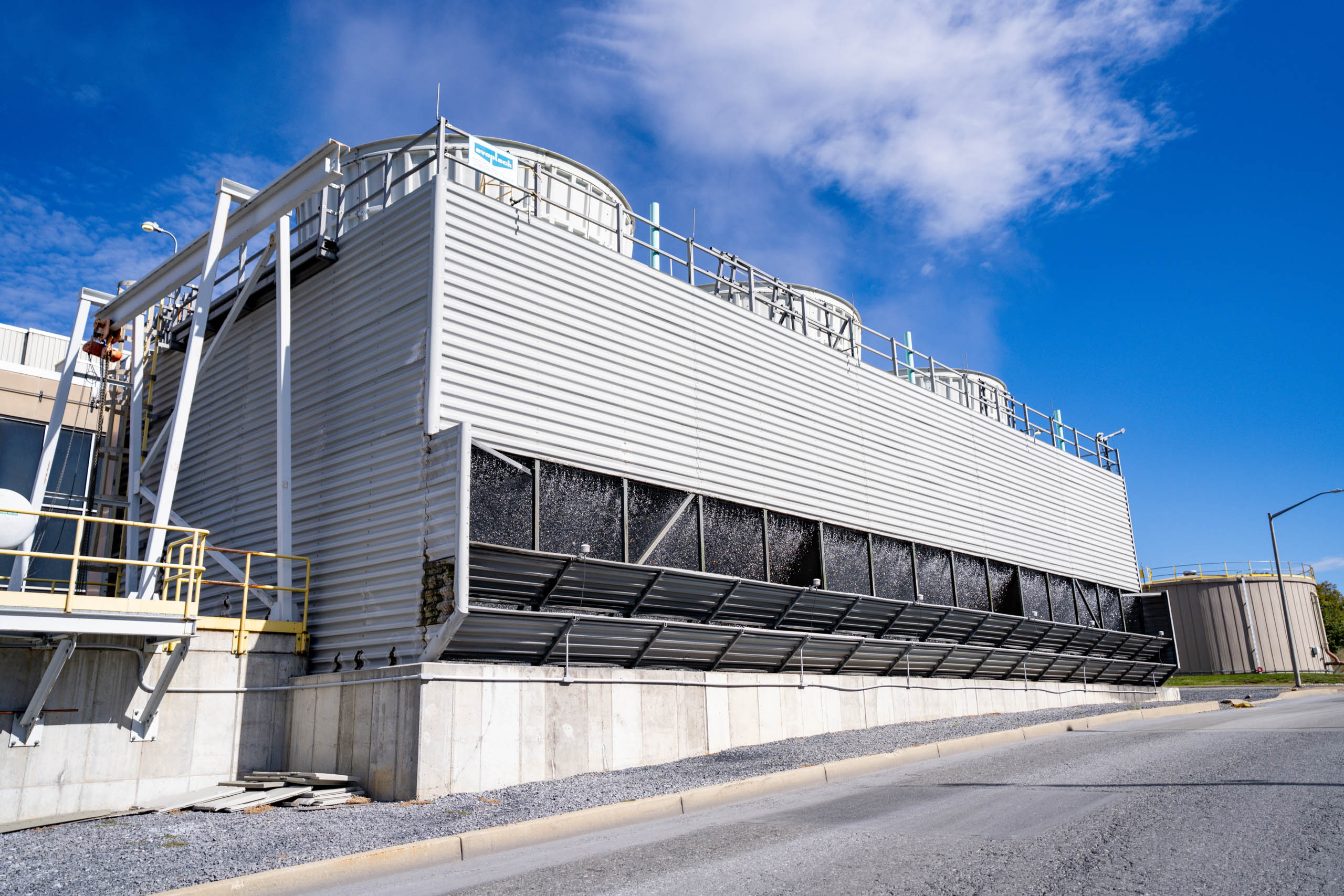 Large lcswma industrial cooling tower with metal panels and ventilation fans on top, located outdoors on a concrete base, with a blue sky and scattered clouds in the background.