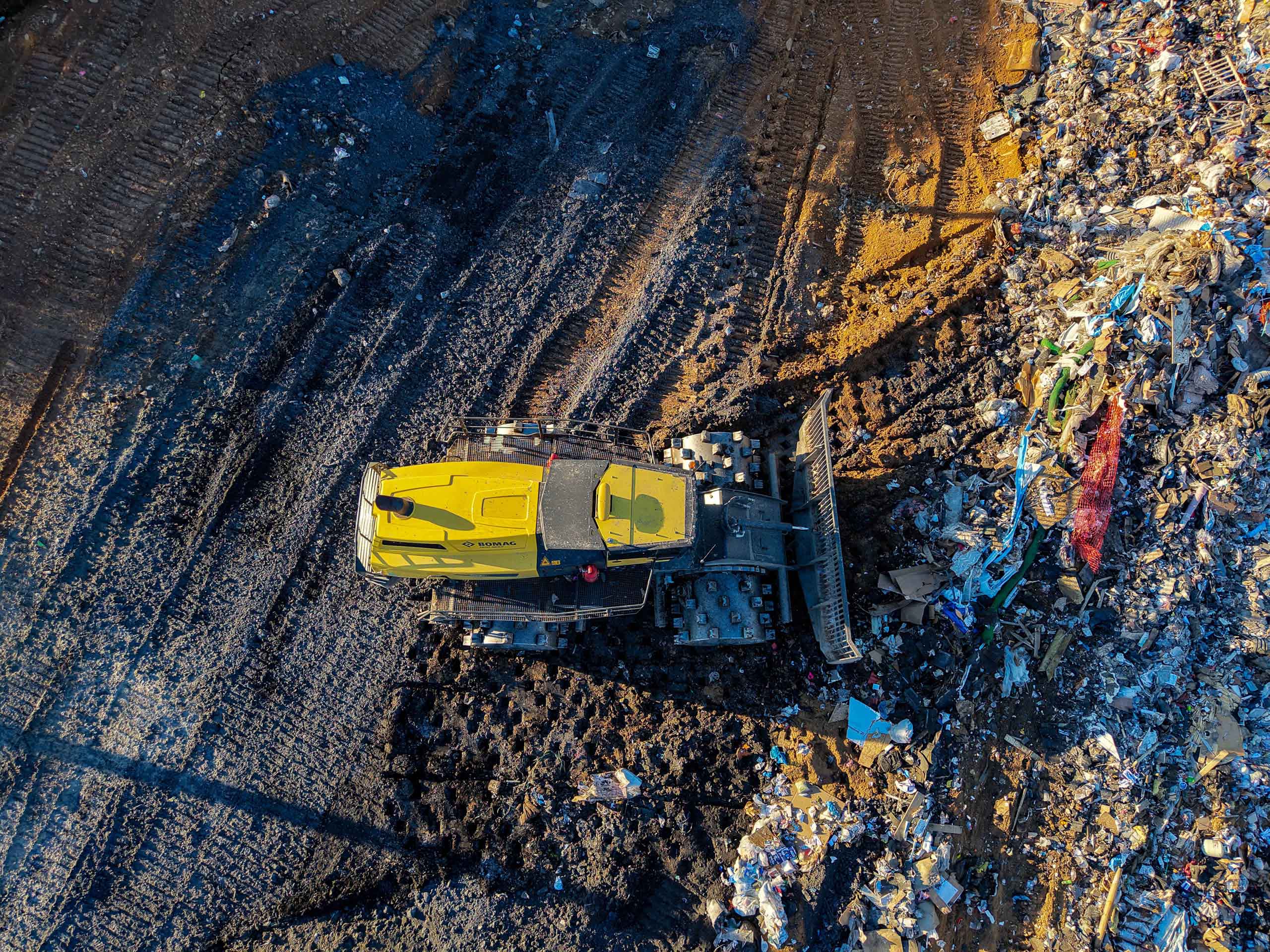 A yellow bulldozer pushes trash and debris at a landfill site, leaving tire tracks in the dirt and surrounded by scattered garbage. The image is taken from an aerial perspective.