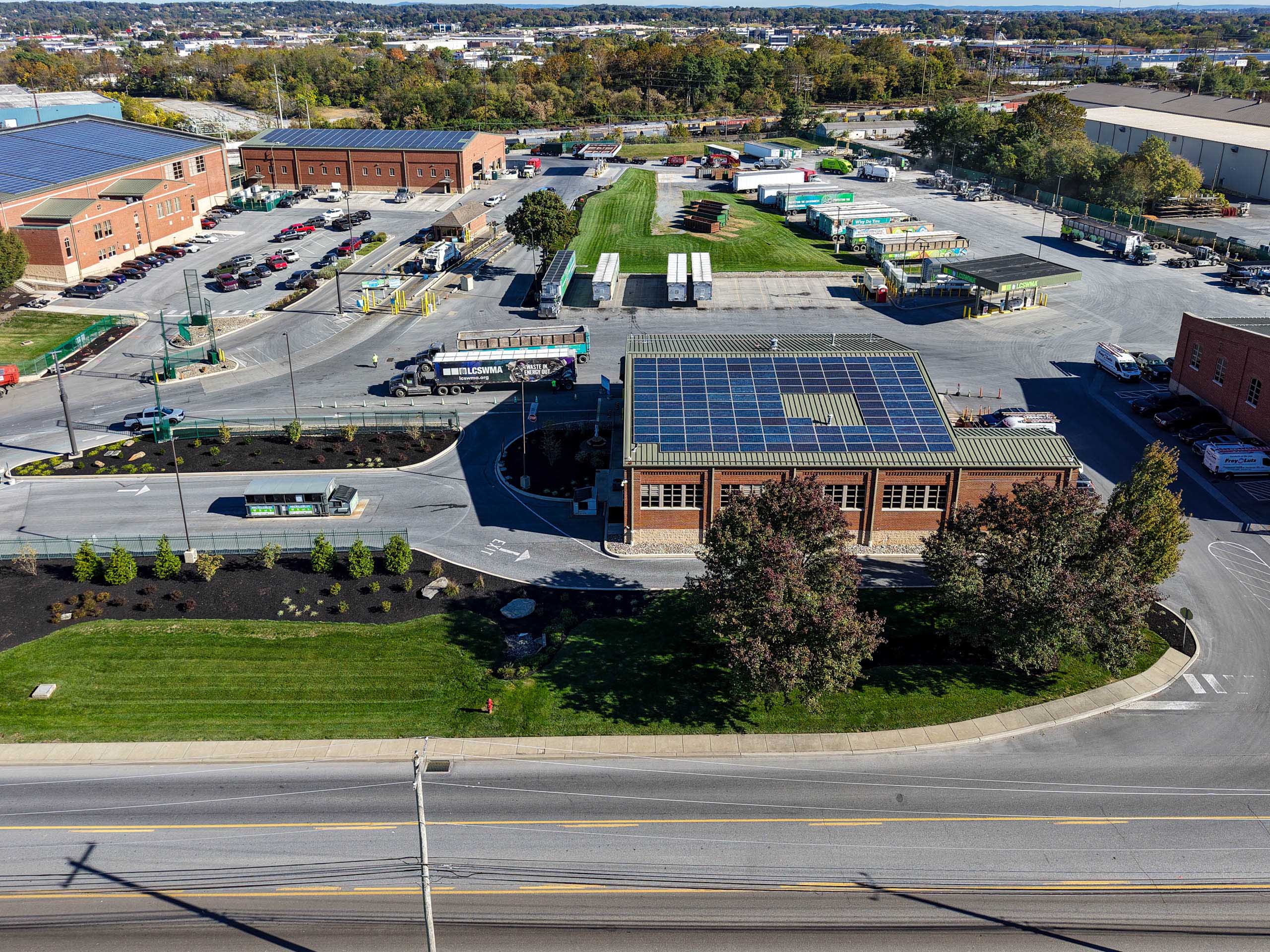 Aerial view of lcswma commercial complex with brick buildings, solar panels, parked trucks, green lawns, and landscaped areas—perfect to Plan Your Drop-off with easy access from the wide road and plenty of surrounding trees.