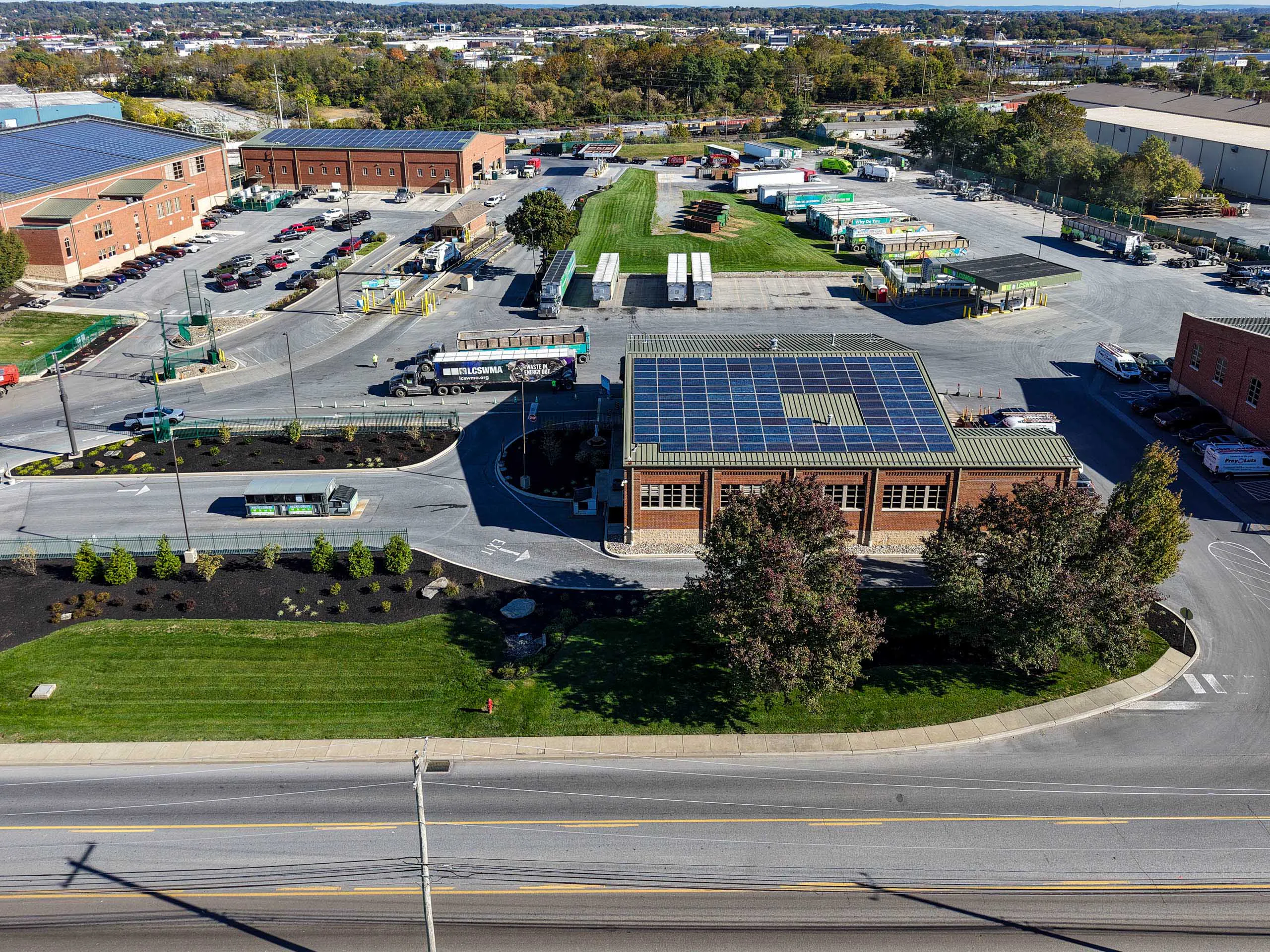 Aerial view of lcswma commercial complex with brick buildings, solar panels, parked trucks, green lawns, and landscaped areas—perfect to Plan Your Drop-off with easy access from the wide road and plenty of surrounding trees.