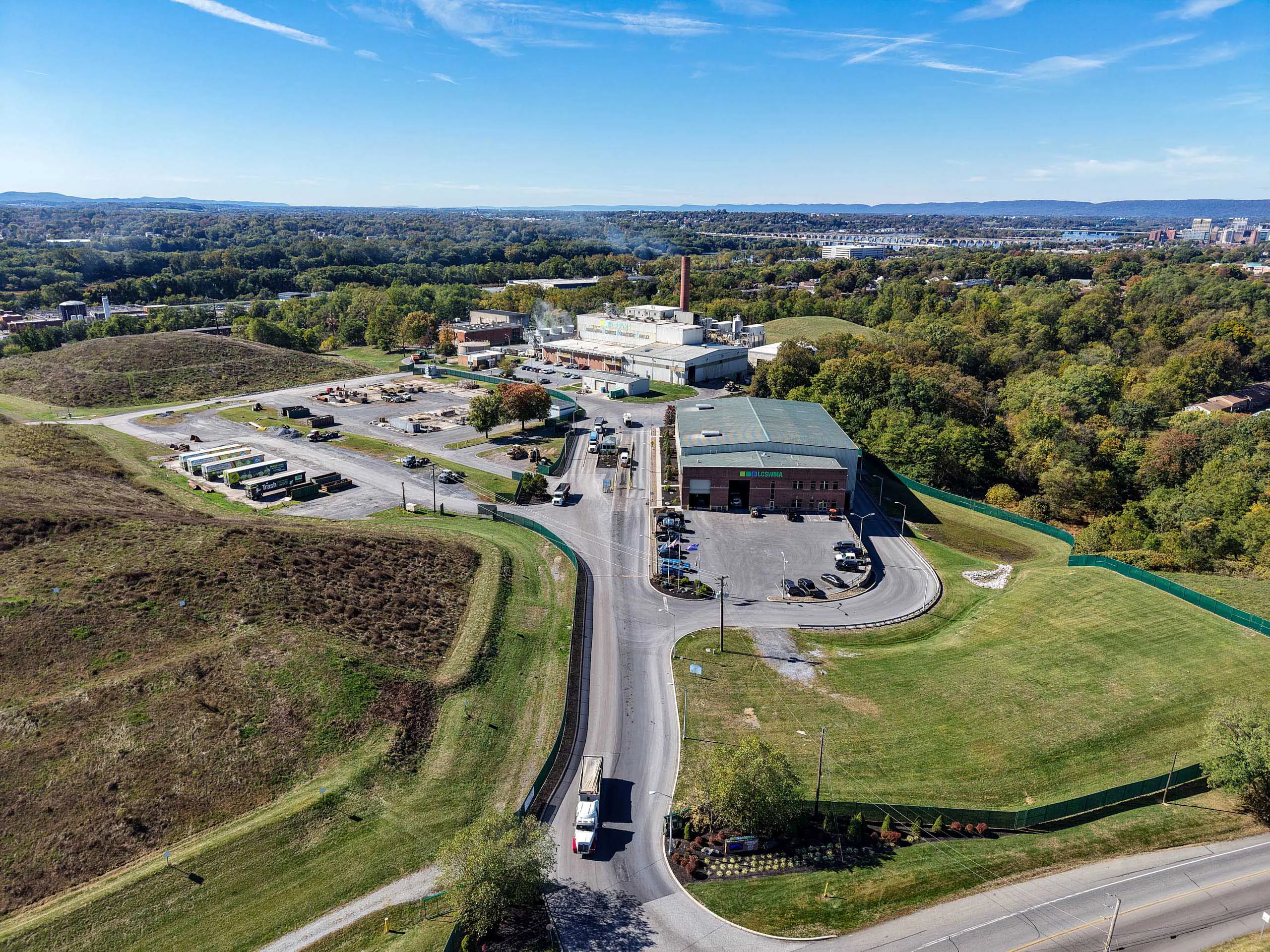 Aerial view of an lcswma facility with several buildings, parking lots, and vehicles, surrounded by grassy areas, trees, and distant hills under a blue sky.