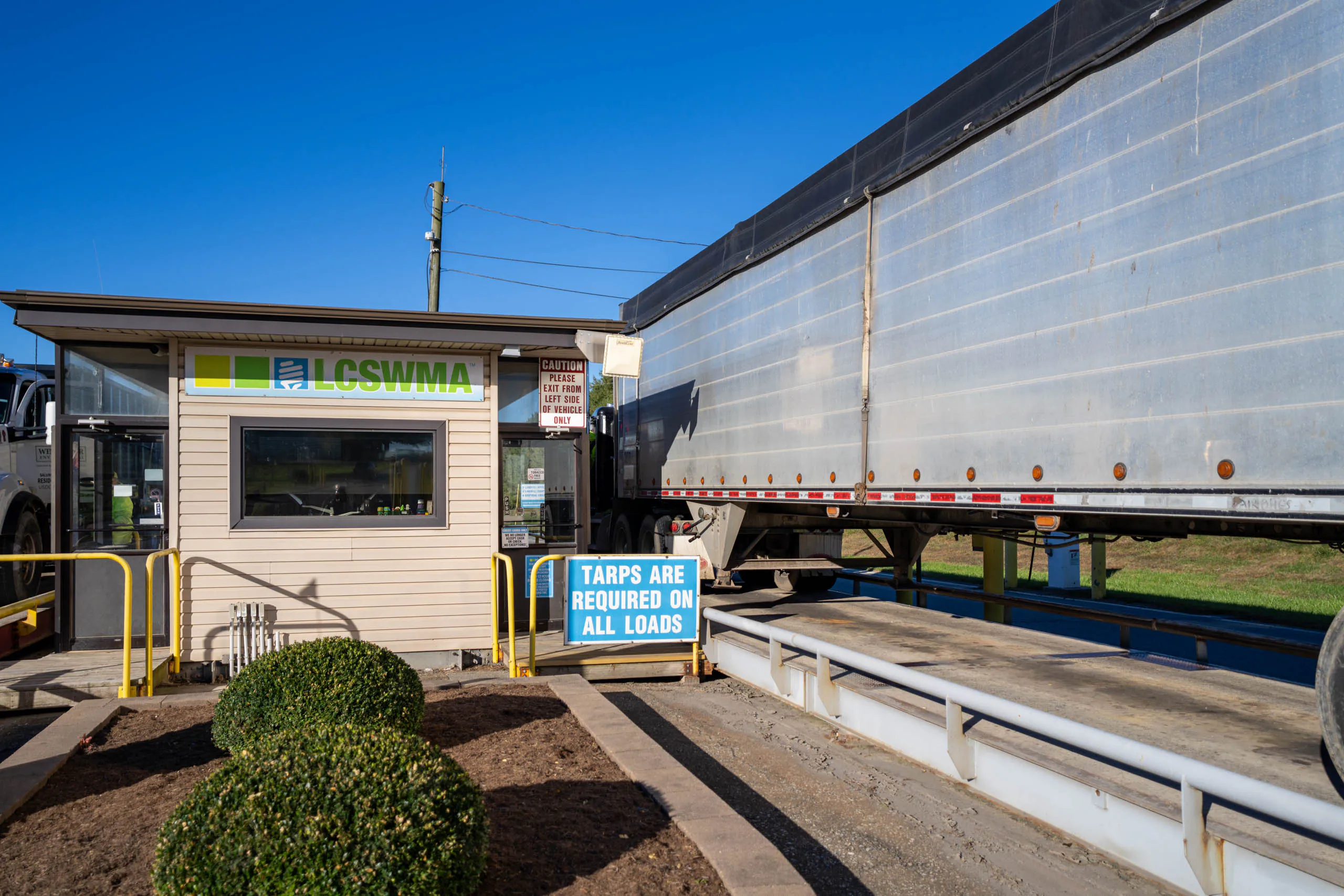 A large truck is parked on a lcswma weigh station scale next to a small building labeled LCSWMA. A blue sign reads, Plan Your Drop-off: Tarps are required on all loads. Bright, clear sky overhead.