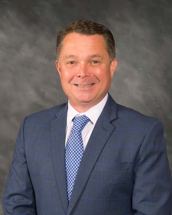 A man in a blue checked suit, white shirt, and blue patterned tie is smiling at the camera against a gray studio background.