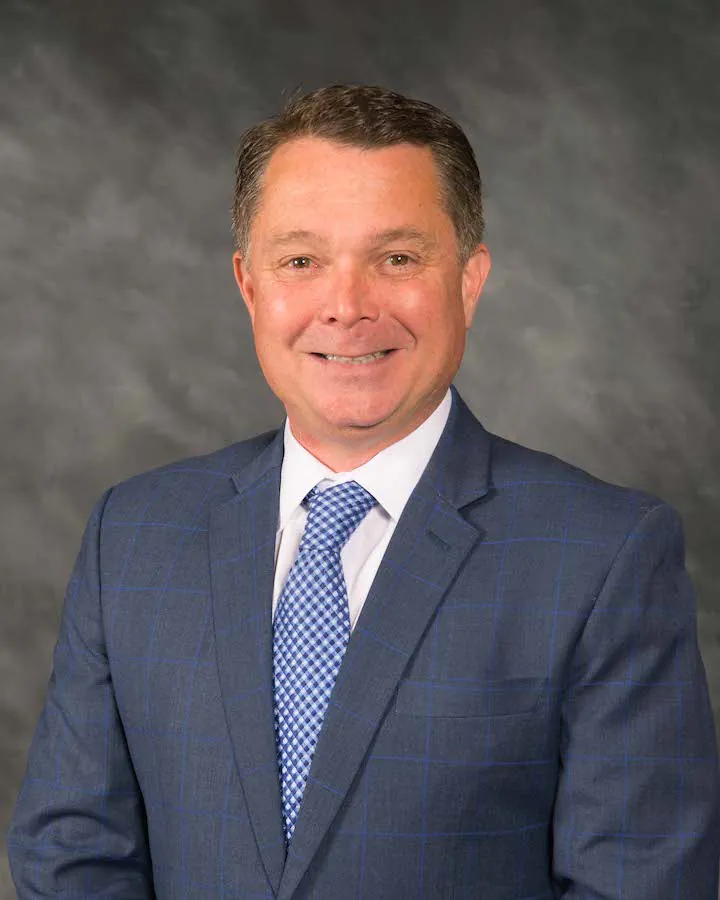 A man in a blue checked suit, white shirt, and blue patterned tie is smiling at the camera against a gray studio background.