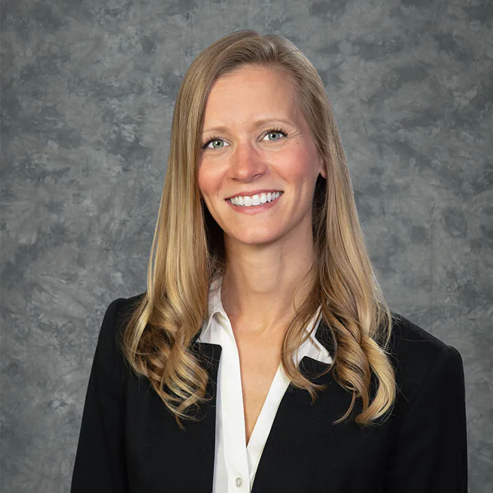 A woman with long blonde hair, wearing a black blazer over a white shirt, smiles at the camera in front of a mottled gray background.