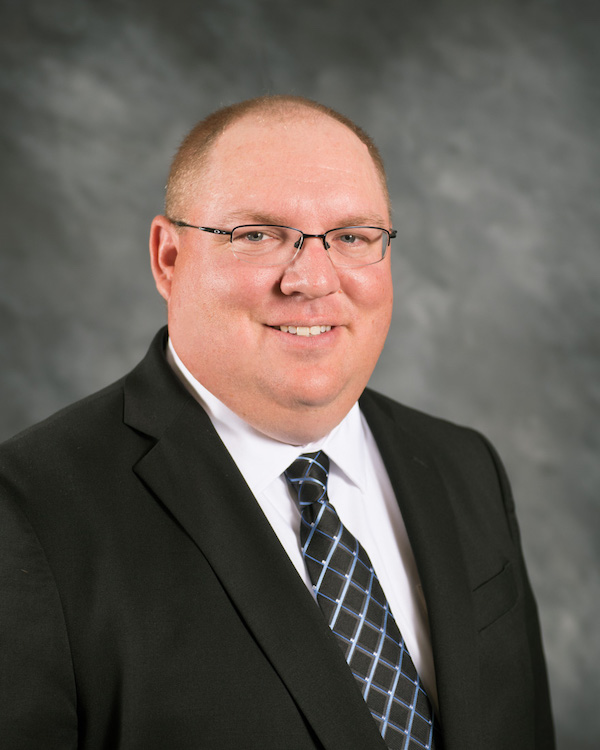 A man wearing glasses, a black suit, white shirt, and patterned tie stands in front of a gray, blurred background, smiling at the camera.