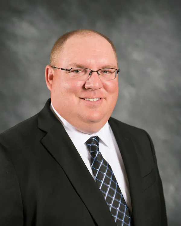A man wearing glasses, a black suit, white shirt, and patterned tie stands in front of a gray, blurred background, smiling at the camera.