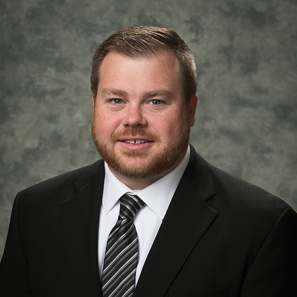 Daniel Youngs, a man with short light brown hair and a beard, wearing a black suit, white shirt, and striped tie, poses in front of a gray mottled background.