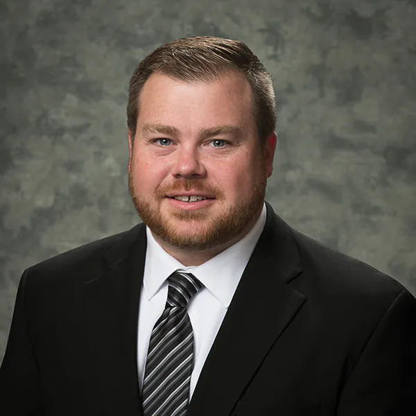 Daniel Youngs, a man with short light brown hair and a beard, wearing a black suit, white shirt, and striped tie, poses in front of a gray mottled background.