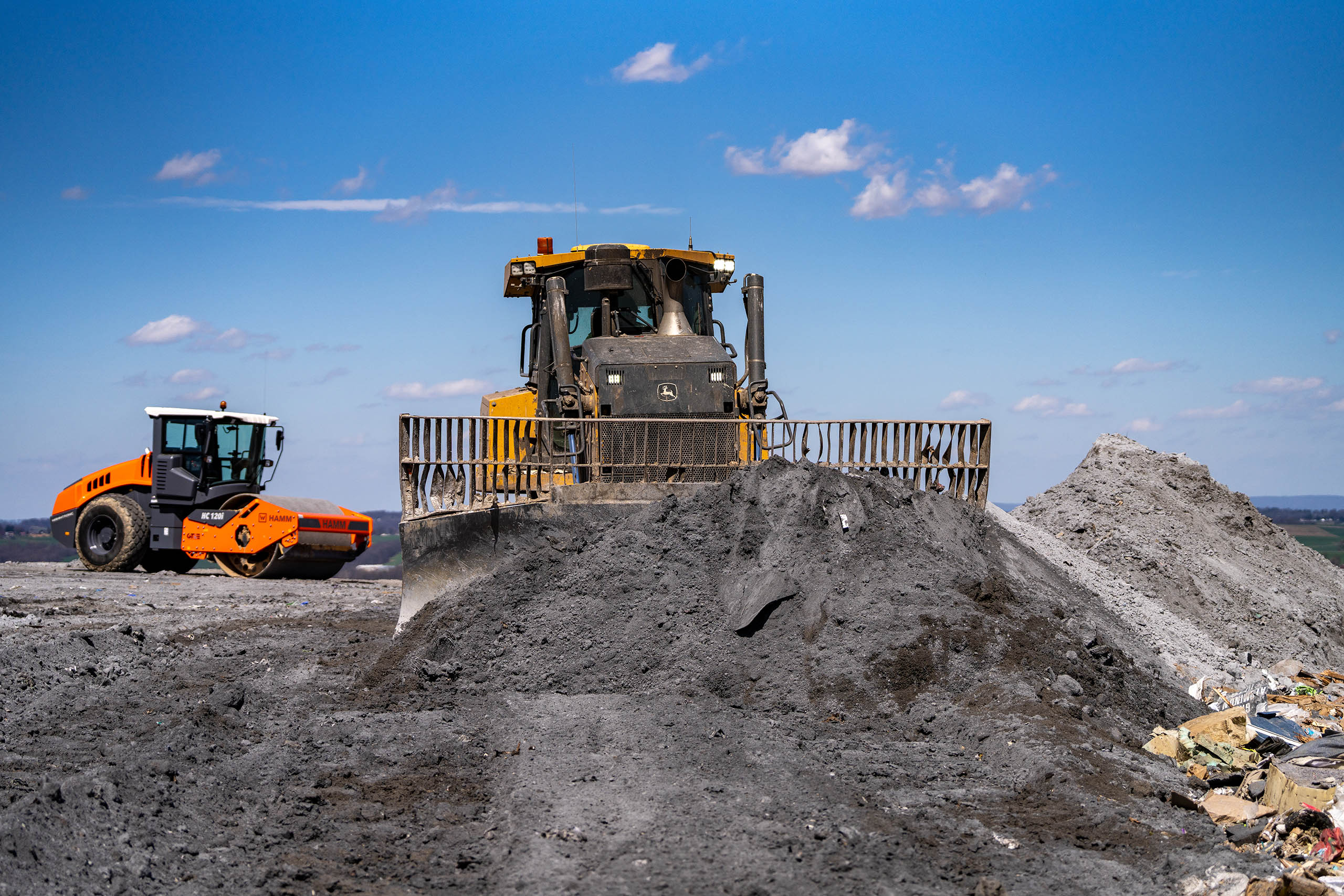 A bulldozer pushes a large pile of dirt at a construction site, while a road roller works in the background under a clear blue sky.