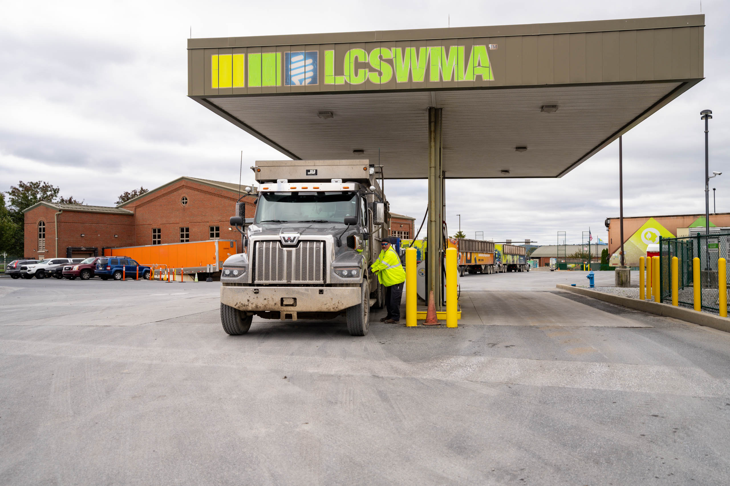 lcswma worker in a yellow jacket assists a large truck at the LCSWMA station, highlighting the partnerships that support operations, with brick buildings and additional vehicles visible in the background under a cloudy sky.