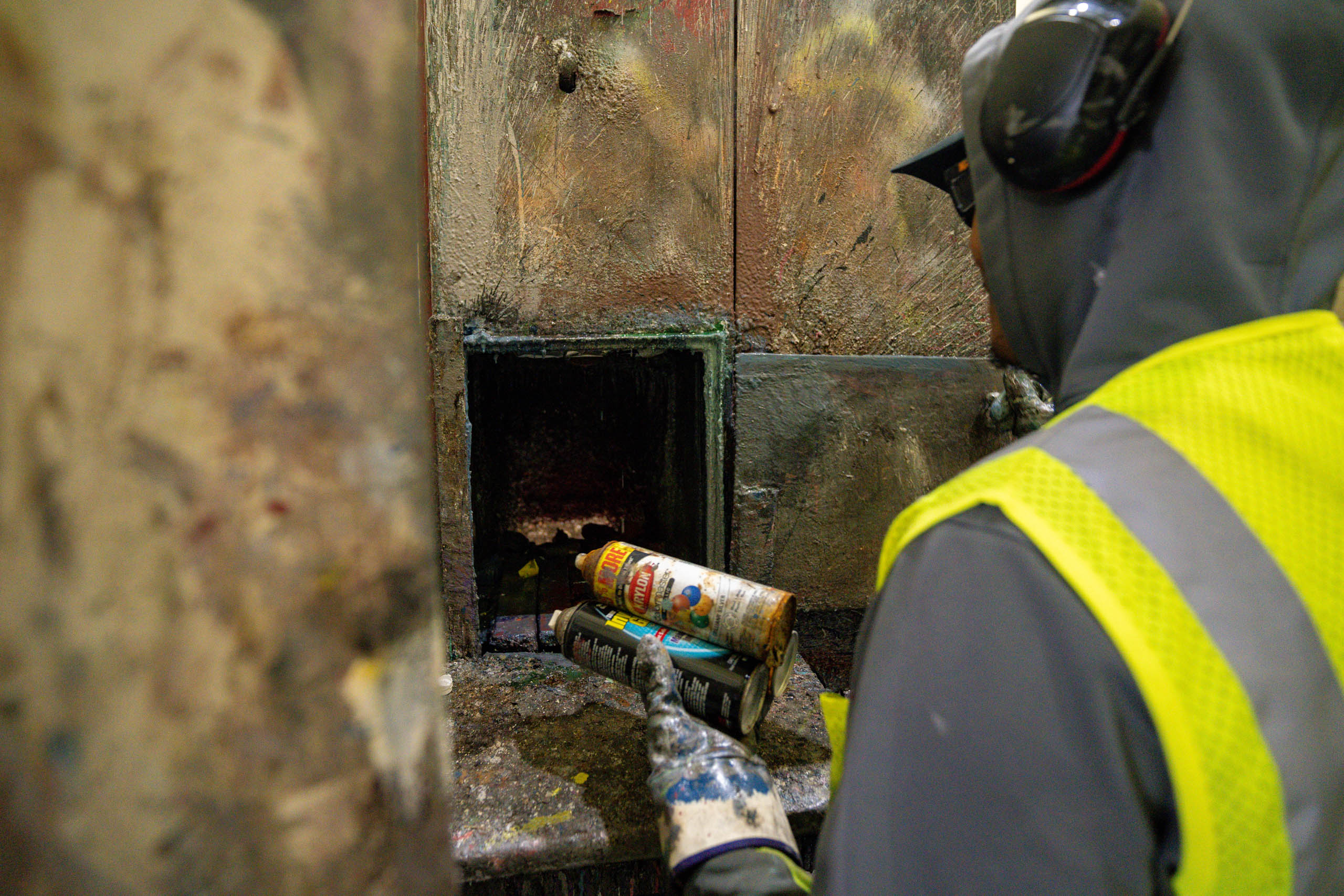 lcswma employee wearing a safety vest, gloves, hoodie, and ear protection, a person disposes of a spray paint can into a small, dirty metal compartment in an industrial setting focused on Green Alternatives.