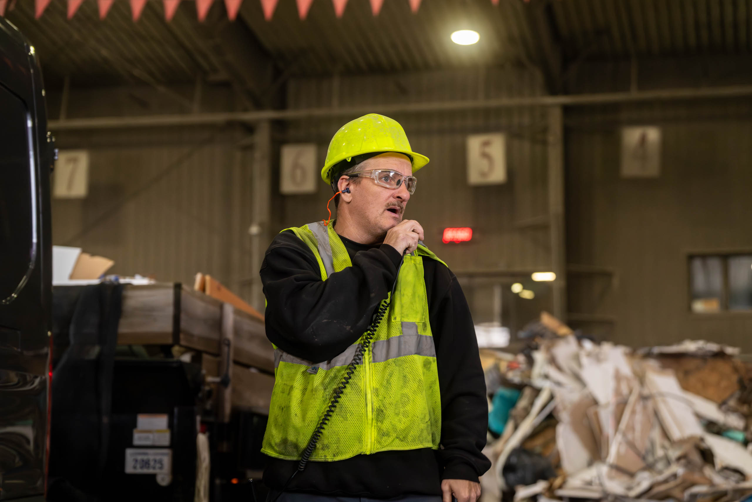 A lcswma worker wearing a yellow hard hat, safety glasses, and a reflective vest speaks into a handheld radio inside an industrial building with debris in the background, coordinating for additional resources.