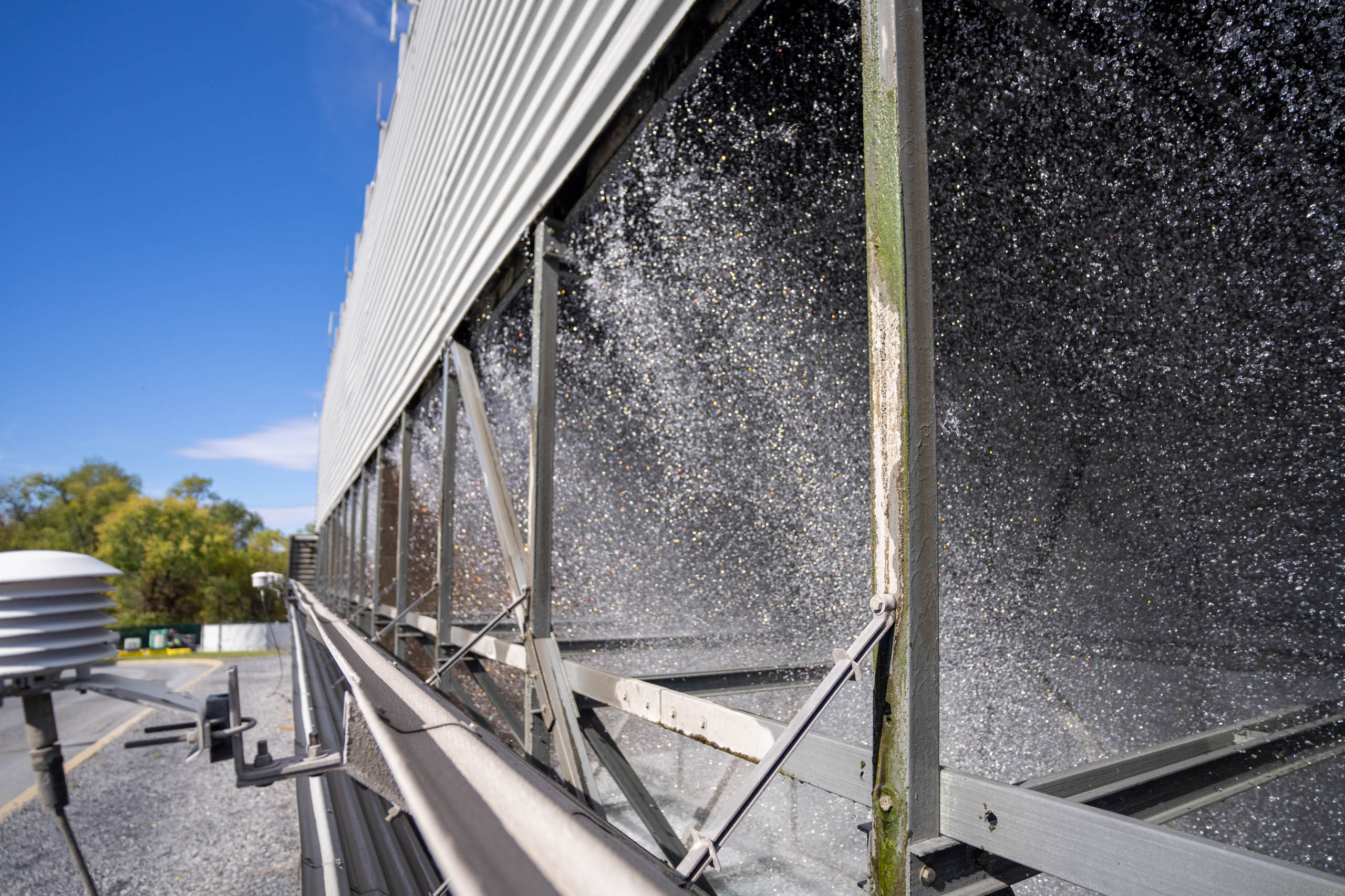 Close-up view of an industrial cooling tower, showing water spraying and cascading inside its metal frame—a testament to advanced cooling programs—with pipes, greenery, and a ventilator in the background under a clear blue sky.
