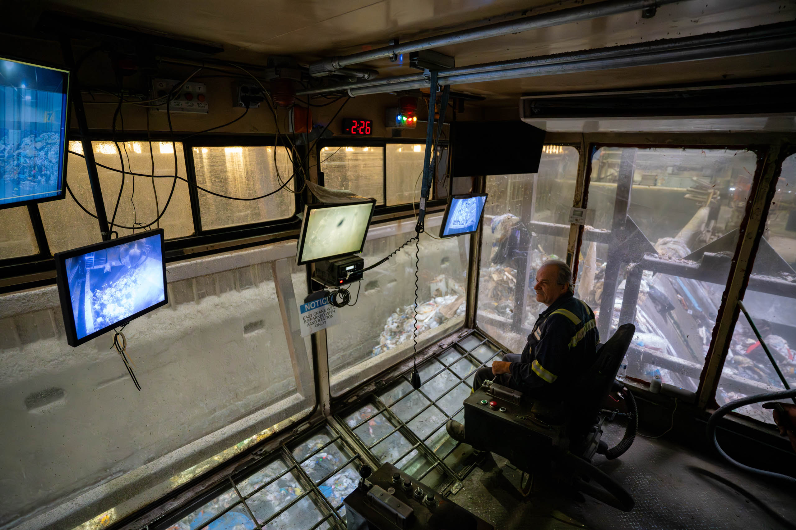 lcswma employee sits in a control room with large windows, monitoring several screens that display images of machinery and materials, overseeing waste tracking operations in an industrial facility filled with debris.