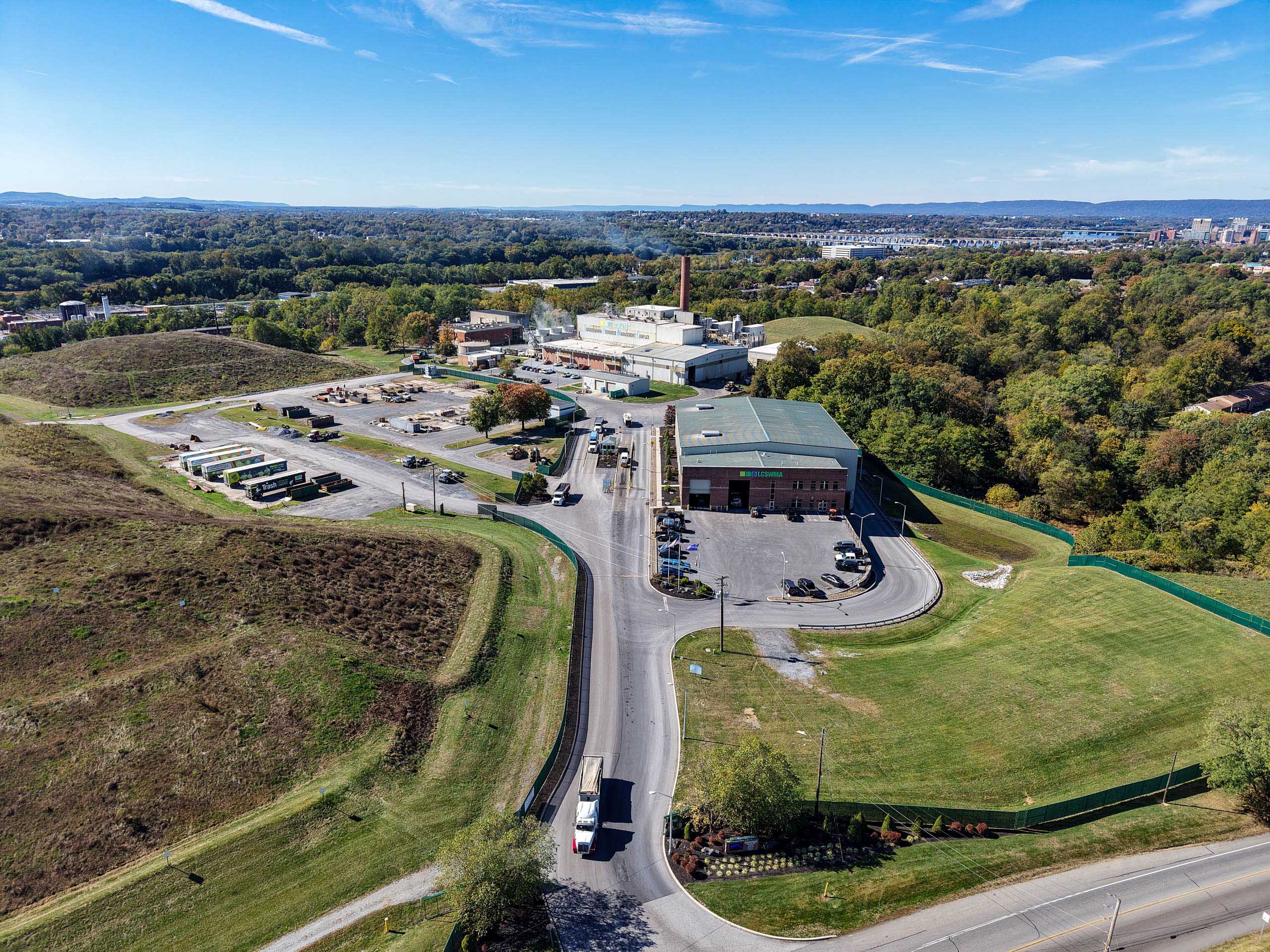 Aerial view of lcswma facility surrounded by grassy hills, trees, and roads, with trucks and equipment visible; the background includes a distant cityscape under a blue sky—ideal for showcasing sites with Additional Resources.