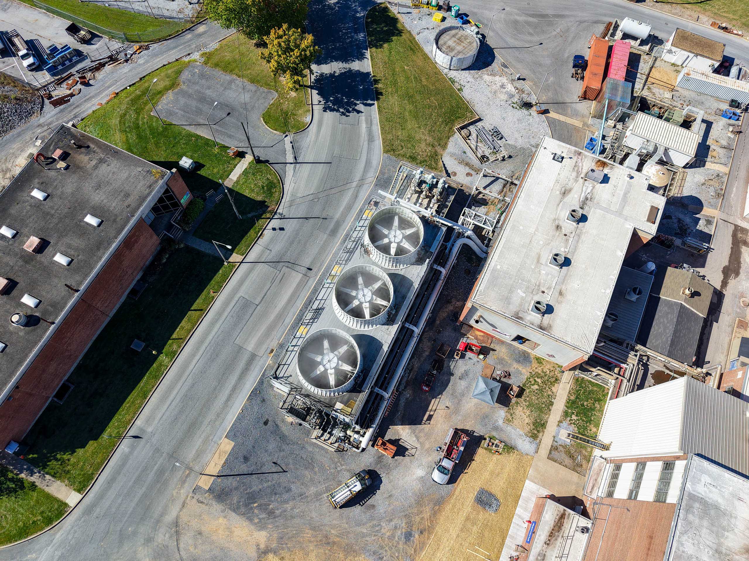 Aerial view of an lcswma facility with large cooling fans, surrounding buildings, parked vehicles, and paved roads intersecting the site. Grassy areas and construction materials are visible nearby.