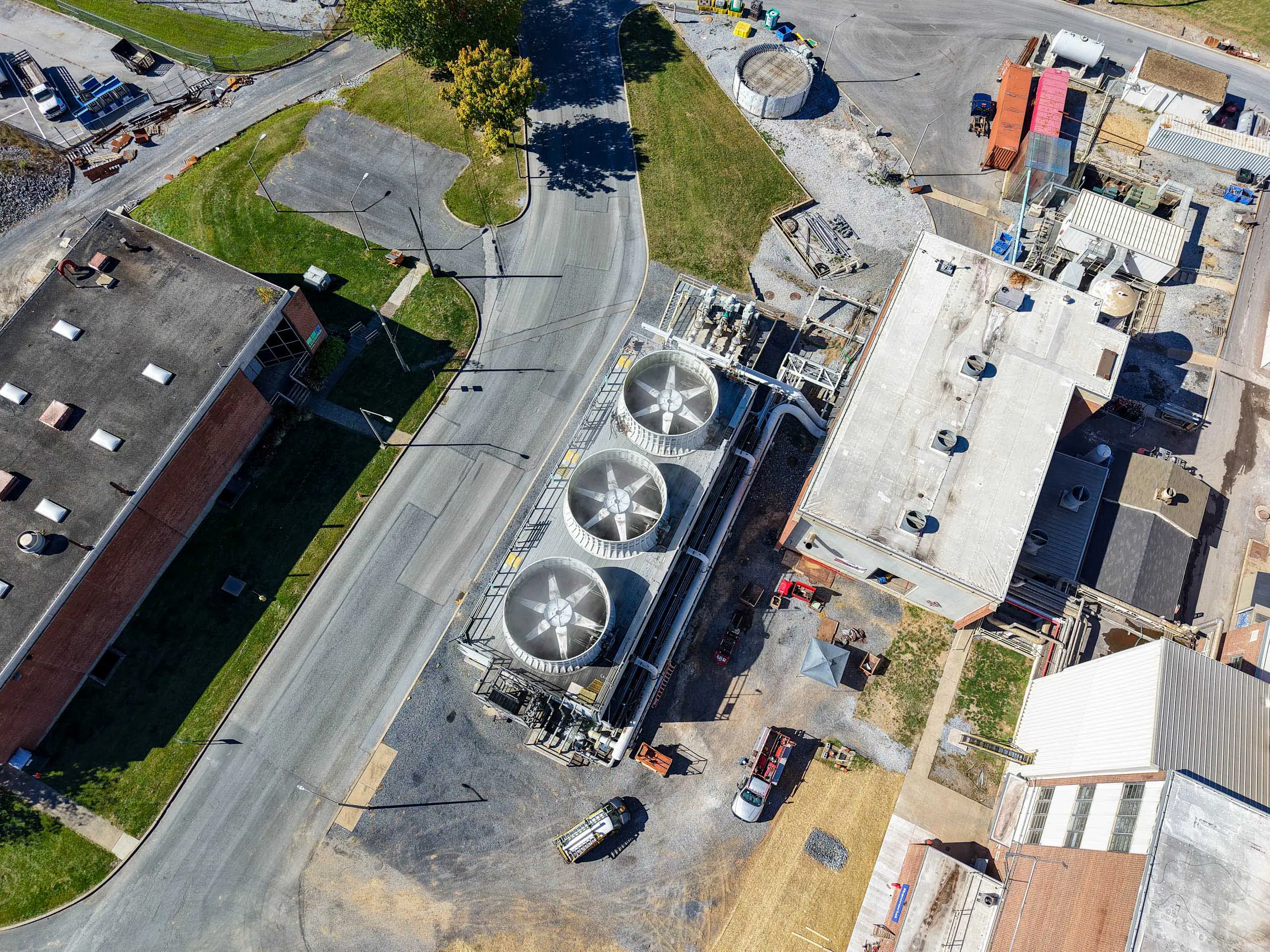 Aerial view of an lcswma facility with large cooling fans, surrounding buildings, parked vehicles, and paved roads intersecting the site. Grassy areas and construction materials are visible nearby.