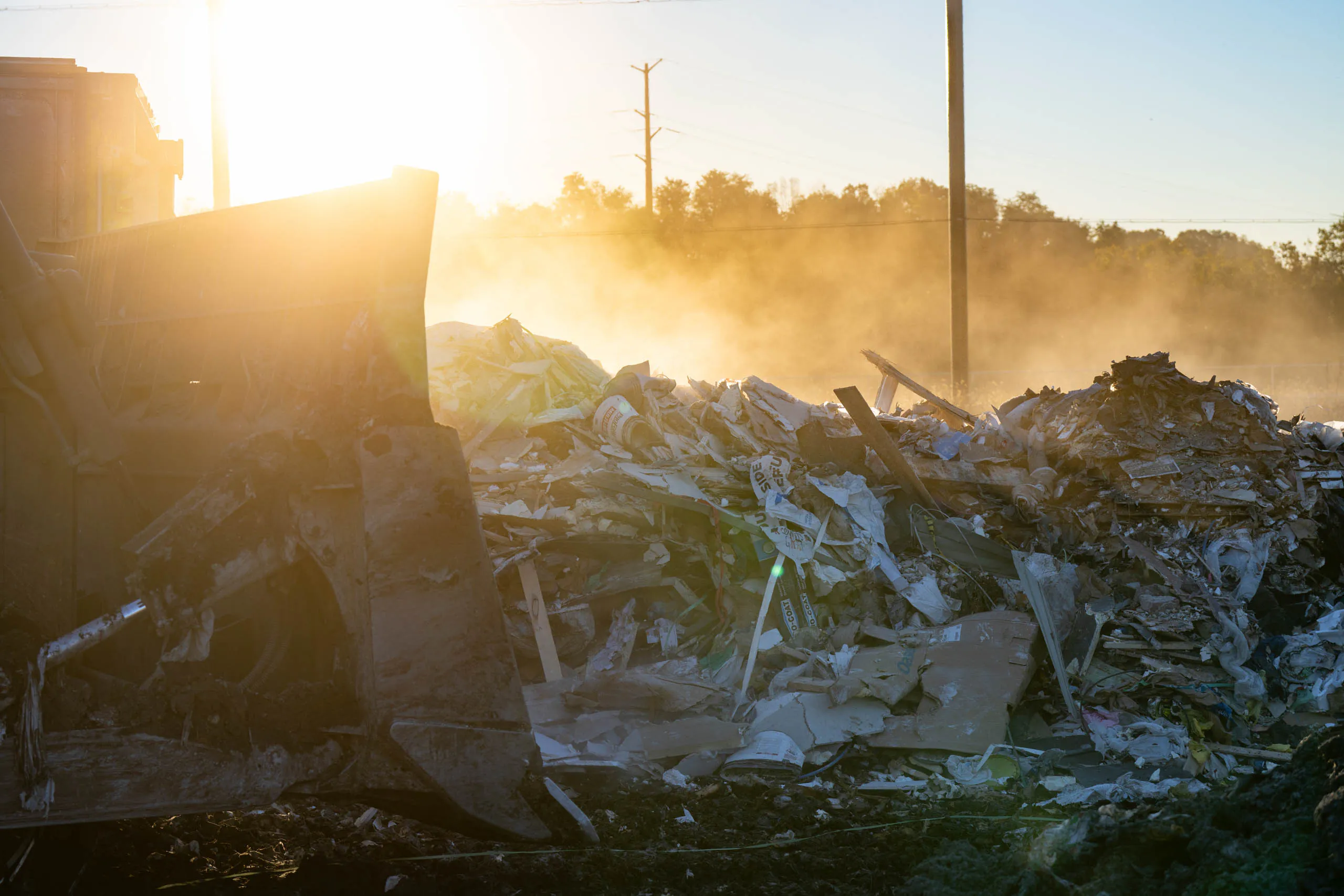 A bulldozer pushes a large pile of debris and garbage at lcswma landfill site, highlighting the growing problem of waste management as the sun shines brightly in the background and dust rises into the air.